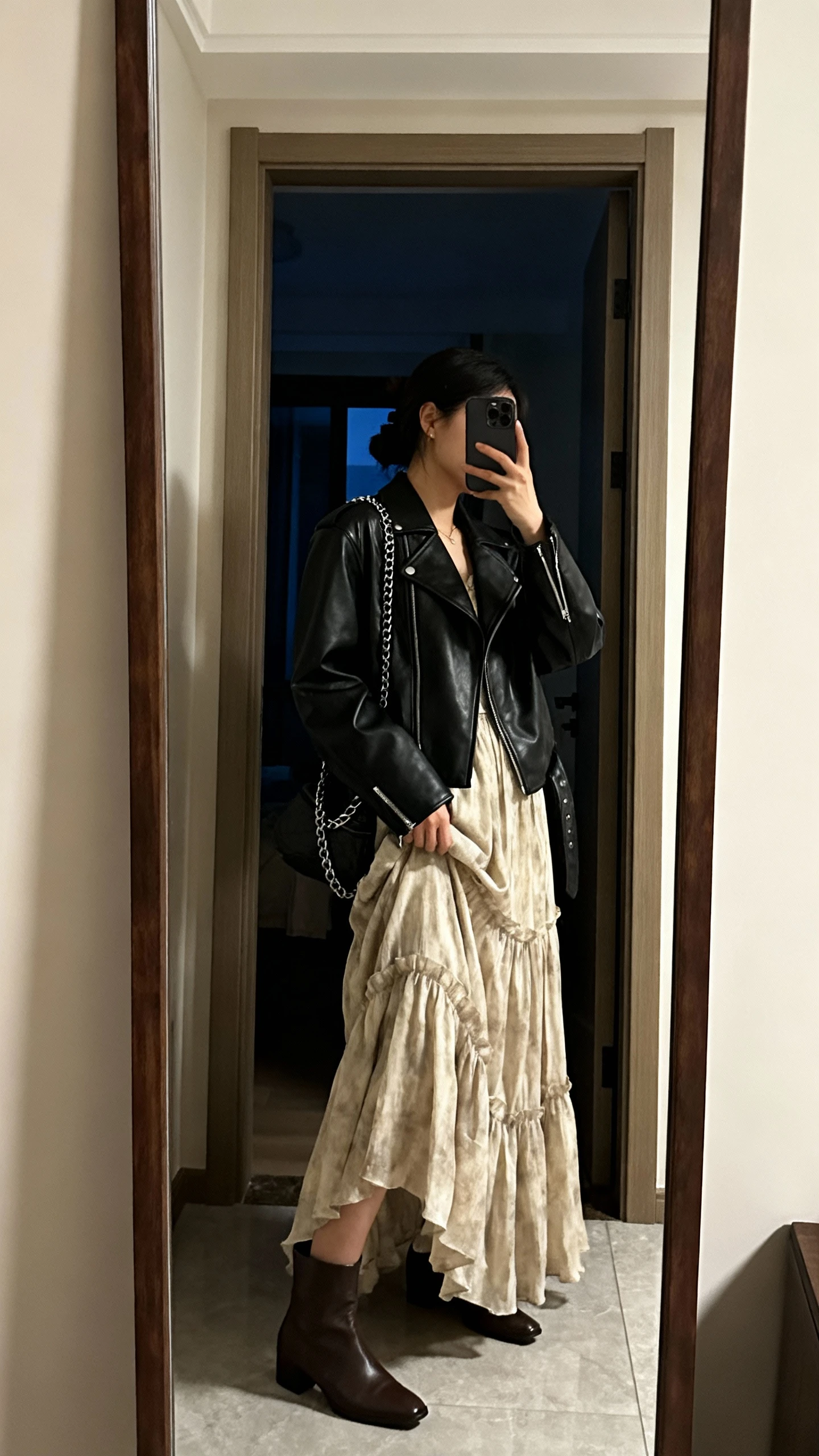 iPhone side-angle mirror selfie of a woman in a flowy maxi dress layered with a black leather jacket, ankle boots, and a chain-strap bag, face not visible, entryway full-length mirror, evening indoor light, iPhone photo quality.