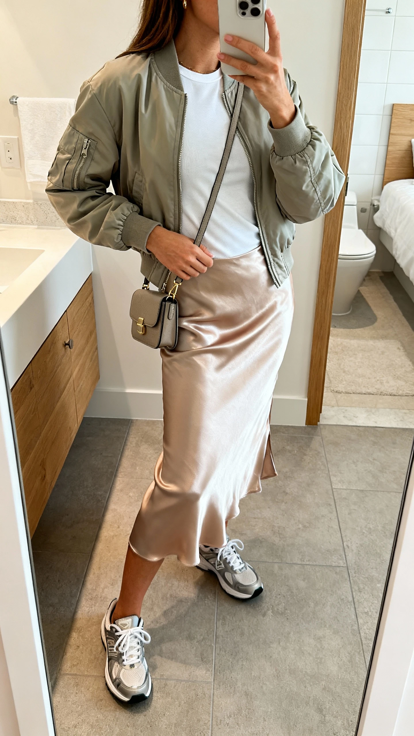 iPhone side-angle mirror selfie of a woman in a lightweight bomber jacket layered over a satin slip skirt with a fitted tee and sleek sneakers, small crossbody bag, face not visible, bathroom mirror with natural indoor light, iPhone photo quality.