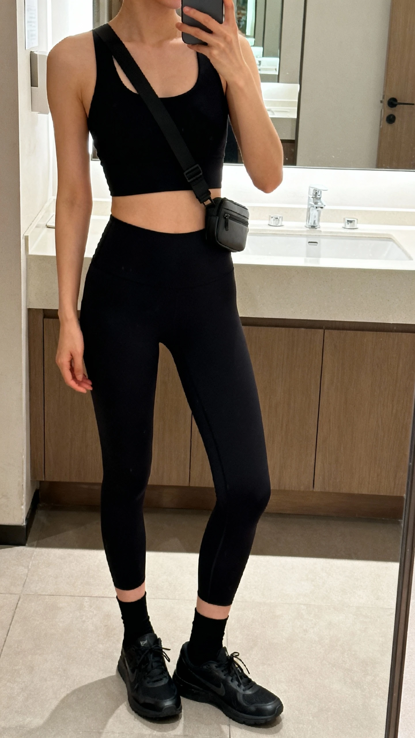 iPhone side-angle mirror selfie of a woman in a monochrome minimalist set: black longline sports bra, black seamless leggings, sleek black running shoes, thin black crew socks, and a minimalist crossbody belt bag, face not visible, cropped at shoulders, bathroom mirror, soft indoor lighting, iPhone photo quality