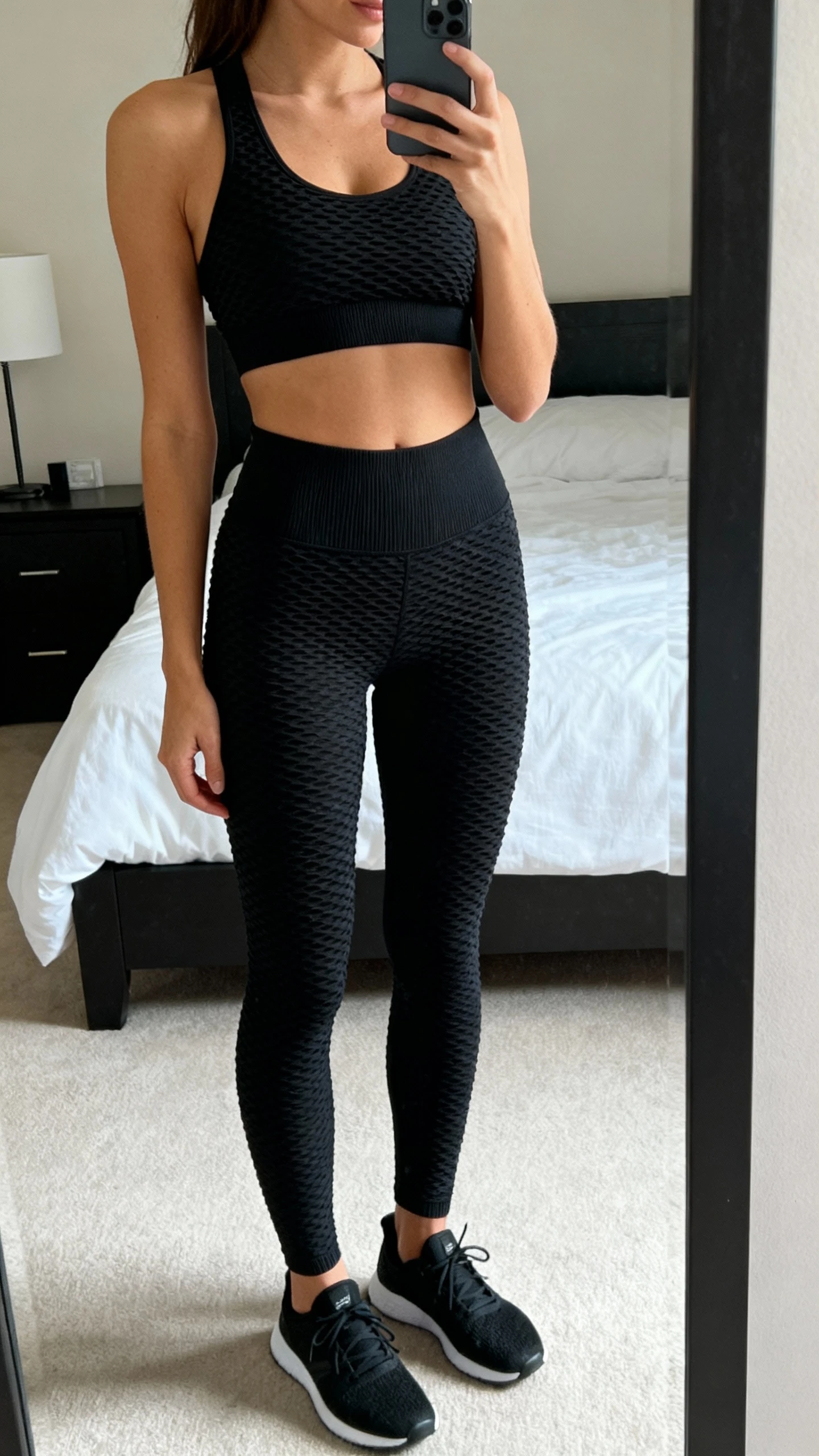 iPhone side-angle mirror selfie of a woman in a monochrome black textured training set (ribbed or jacquard black sports bra and matching high-waist leggings) with black trainers, face not visible, cropped at shoulders, bedroom mirror, soft daylight, iPhone photo quality.