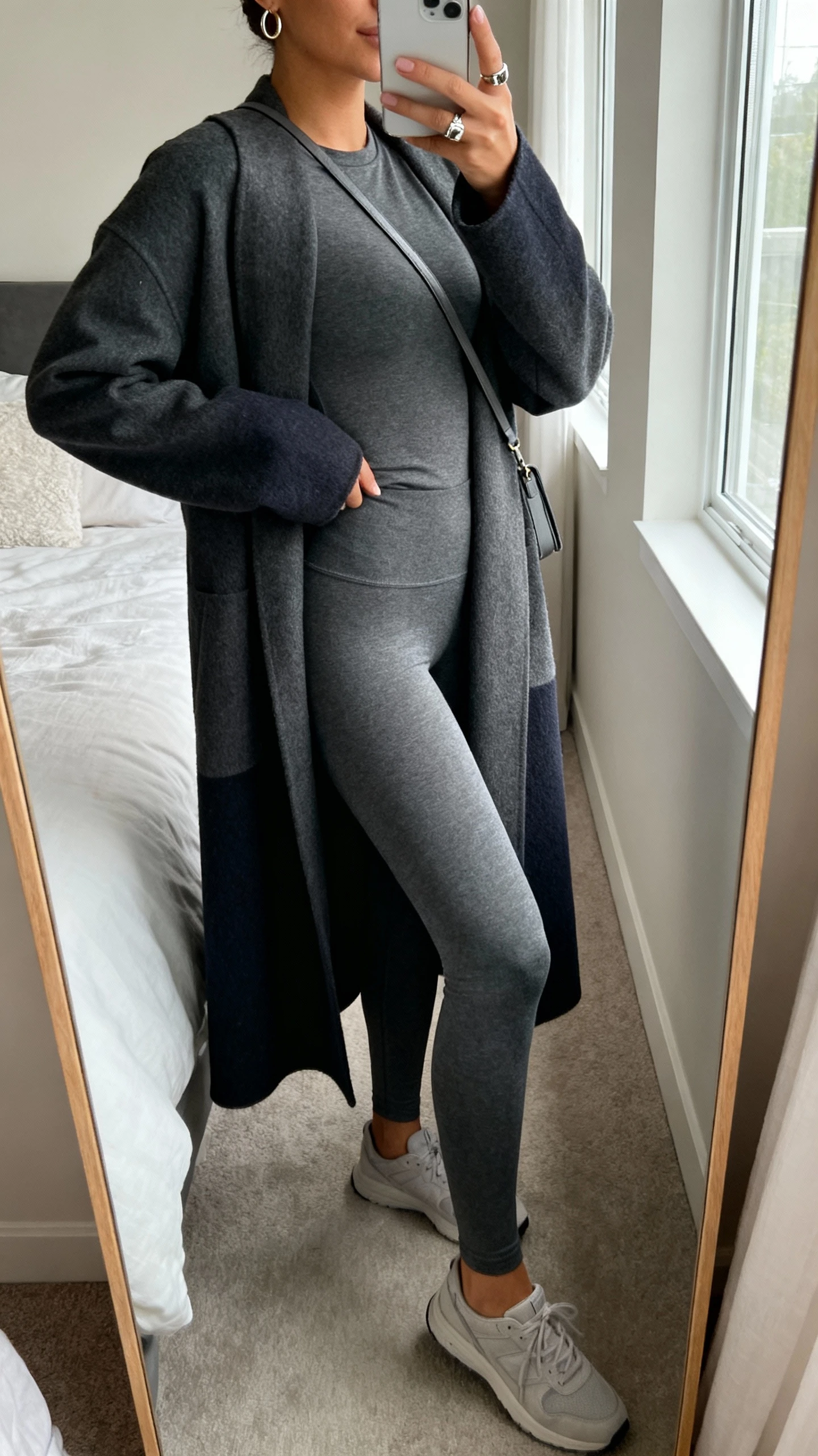 iPhone side-angle mirror selfie of a woman in a monochrome charcoal matching set (leggings and longline fitted top) with a slightly deeper charcoal longline coat, minimalist sneakers, a slim crossbody, small hoop earrings and one statement ring, face not visible, cropped at neck, bedroom full-length mirror, natural window light, iPhone photo quality.