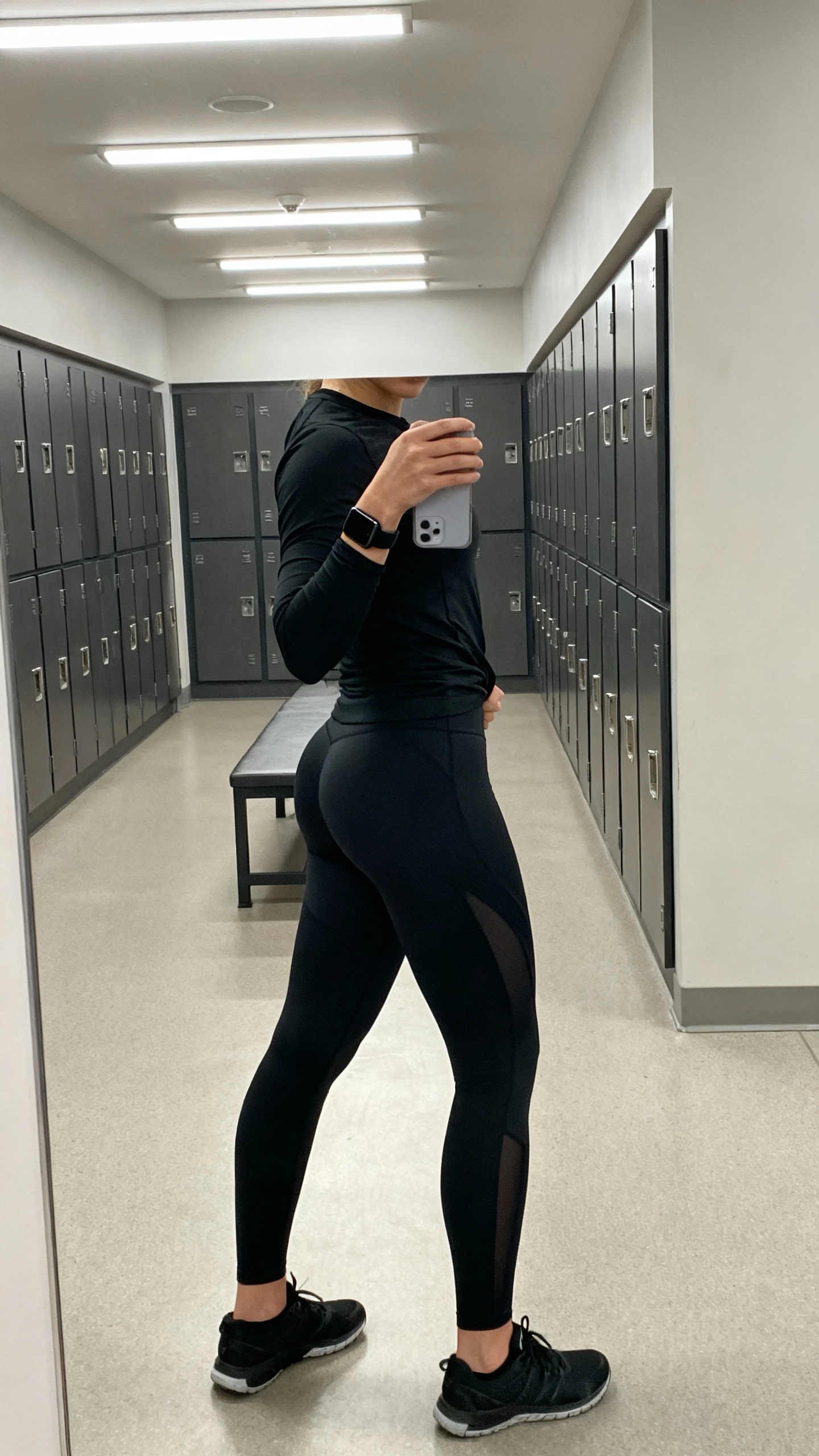 iPhone side-angle mirror selfie of a woman in a performance black long-sleeve top and contoured black leggings, face not visible, gym locker-room mirror with overhead lighting, black running shoes and a slim black armband, iPhone photo quality