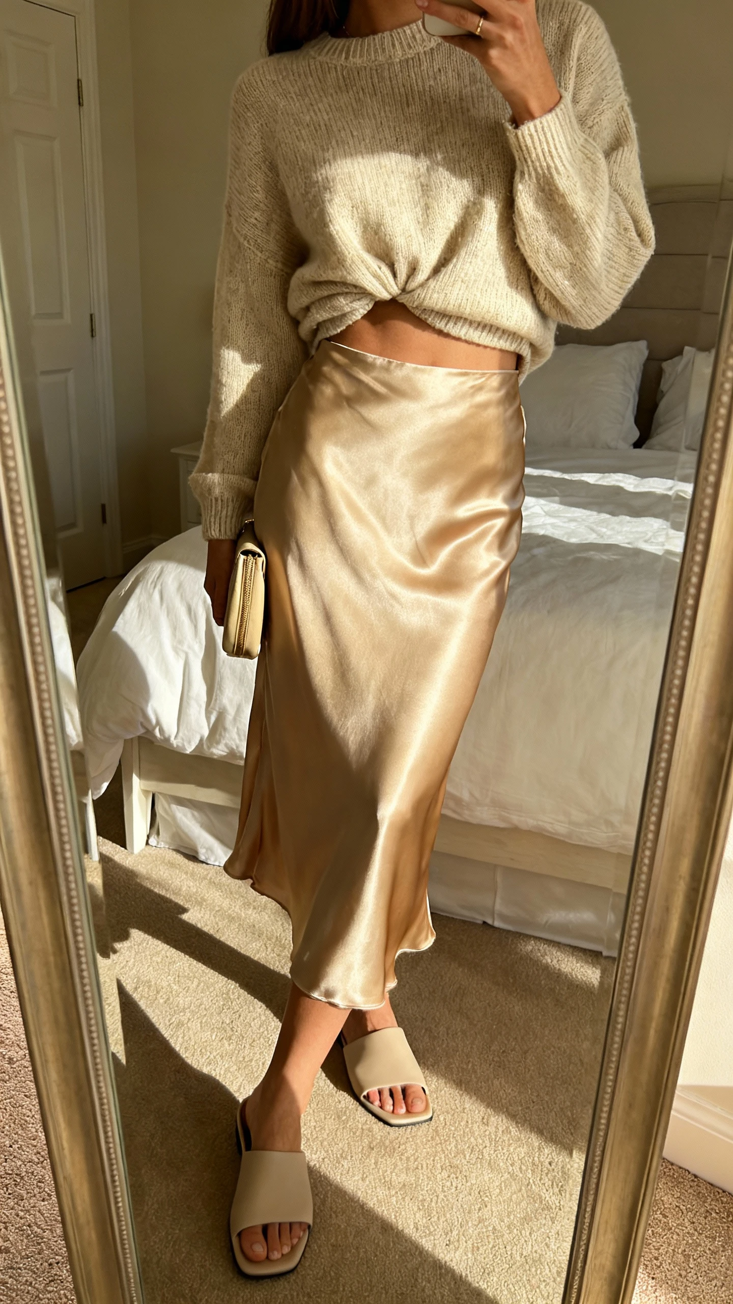 iPhone side-angle mirror selfie of a woman in a satin slip skirt with a lightweight knit sweater half-tucked, minimalist mules, and a small clutch, face not visible, bedroom mirror, gentle morning light, iPhone photo quality.