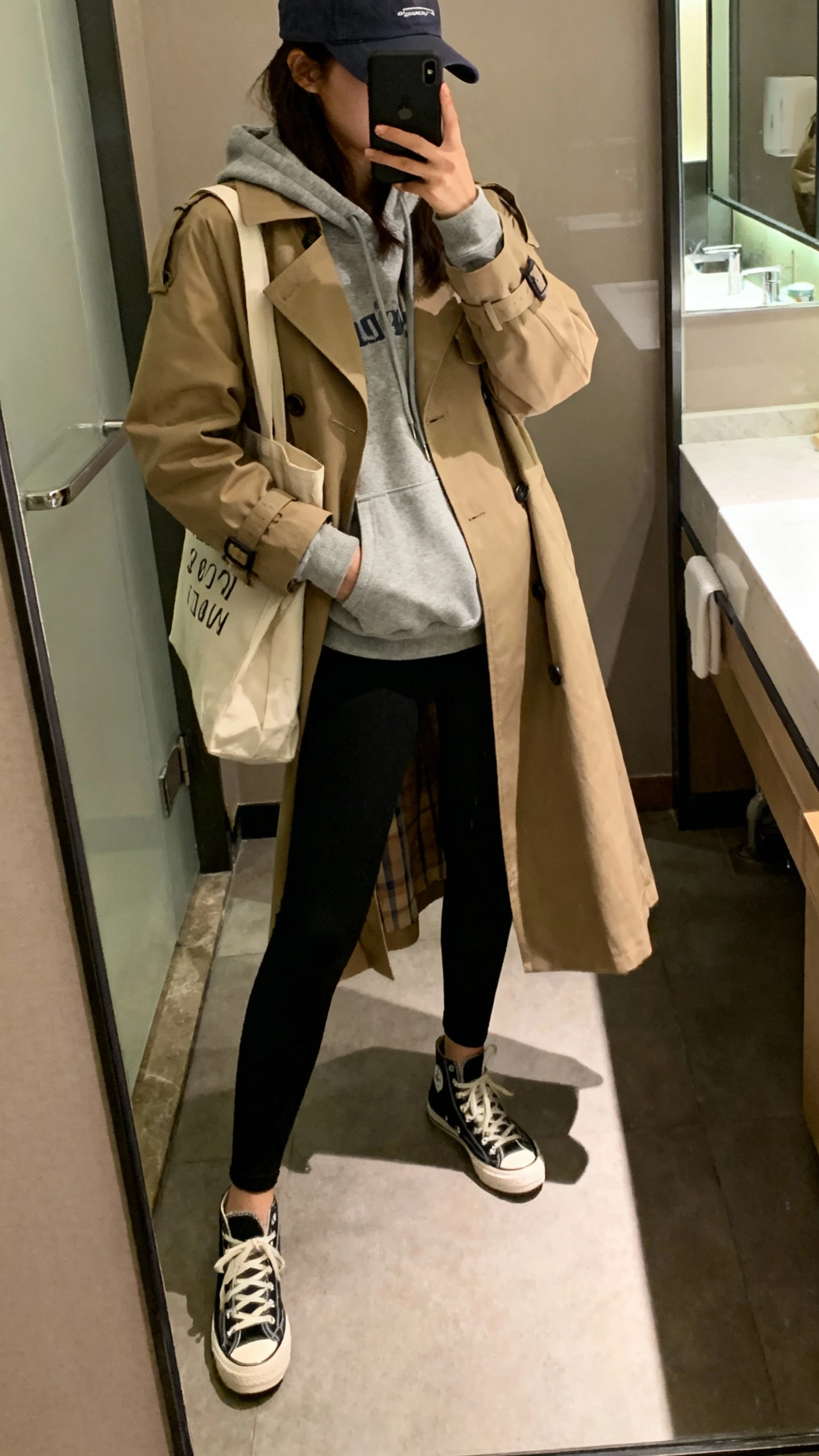 iPhone side-angle mirror selfie of a woman in a street-style hoodie layered under a classic trench coat, black leggings, retro sneakers, tote bag, baseball cap, face not visible, bathroom mirror, indoor lighting, iPhone photo quality.