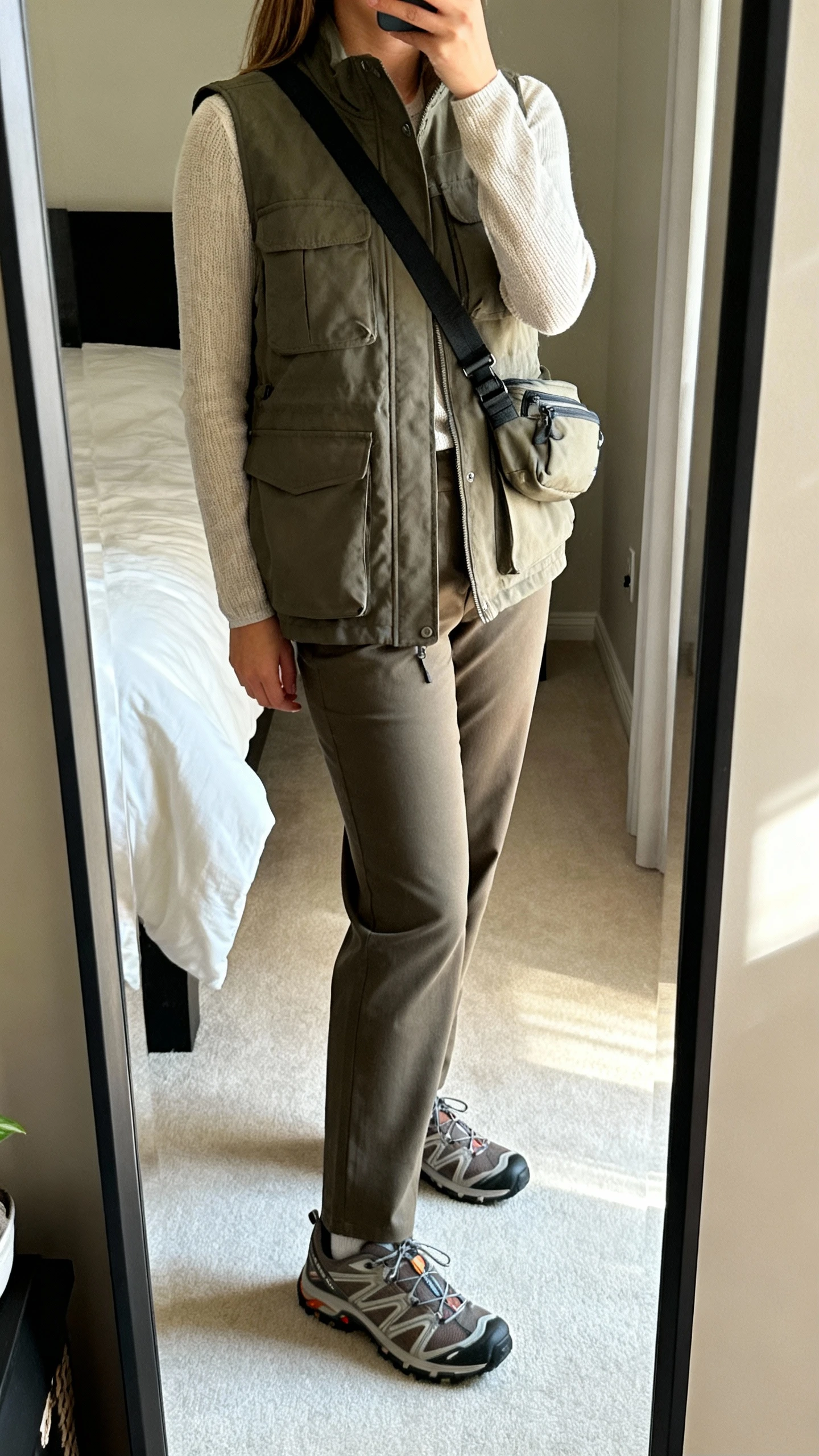 iPhone side-angle mirror selfie of a woman in a utility vest layered over a lightweight knit top with straight-leg trousers and trail-style sneakers, crossbody or belt bag, face not visible, bedroom mirror with soft morning light, iPhone photo quality.