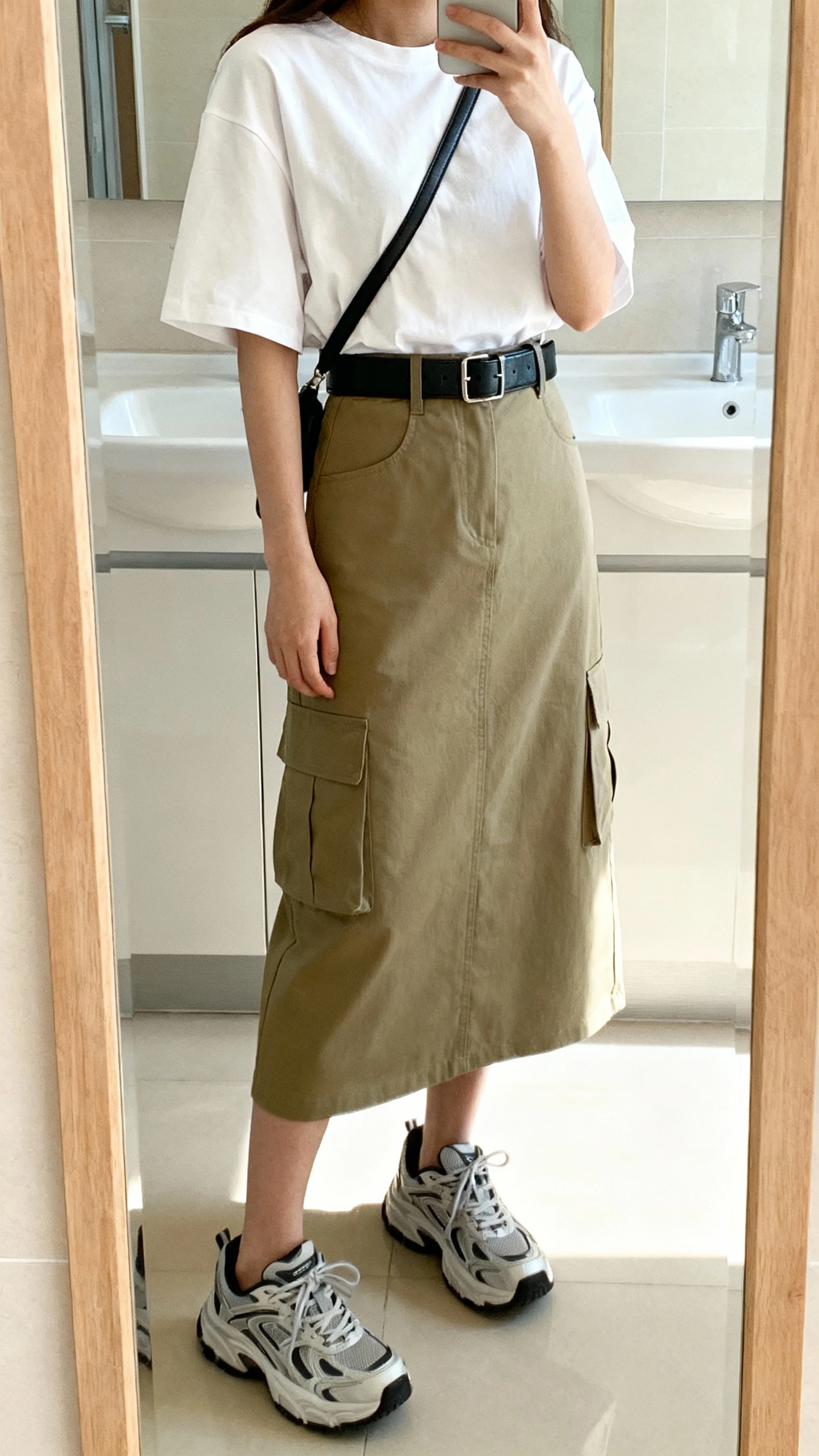 iPhone side-angle mirror selfie of a woman in a utility-chic look: khaki cargo midi skirt, boxy white tee tucked in, black leather belt, chunky sneakers, minimalist crossbody, face not visible, cropped at shoulders, bathroom mirror, bright indoor lighting, iPhone photo quality.