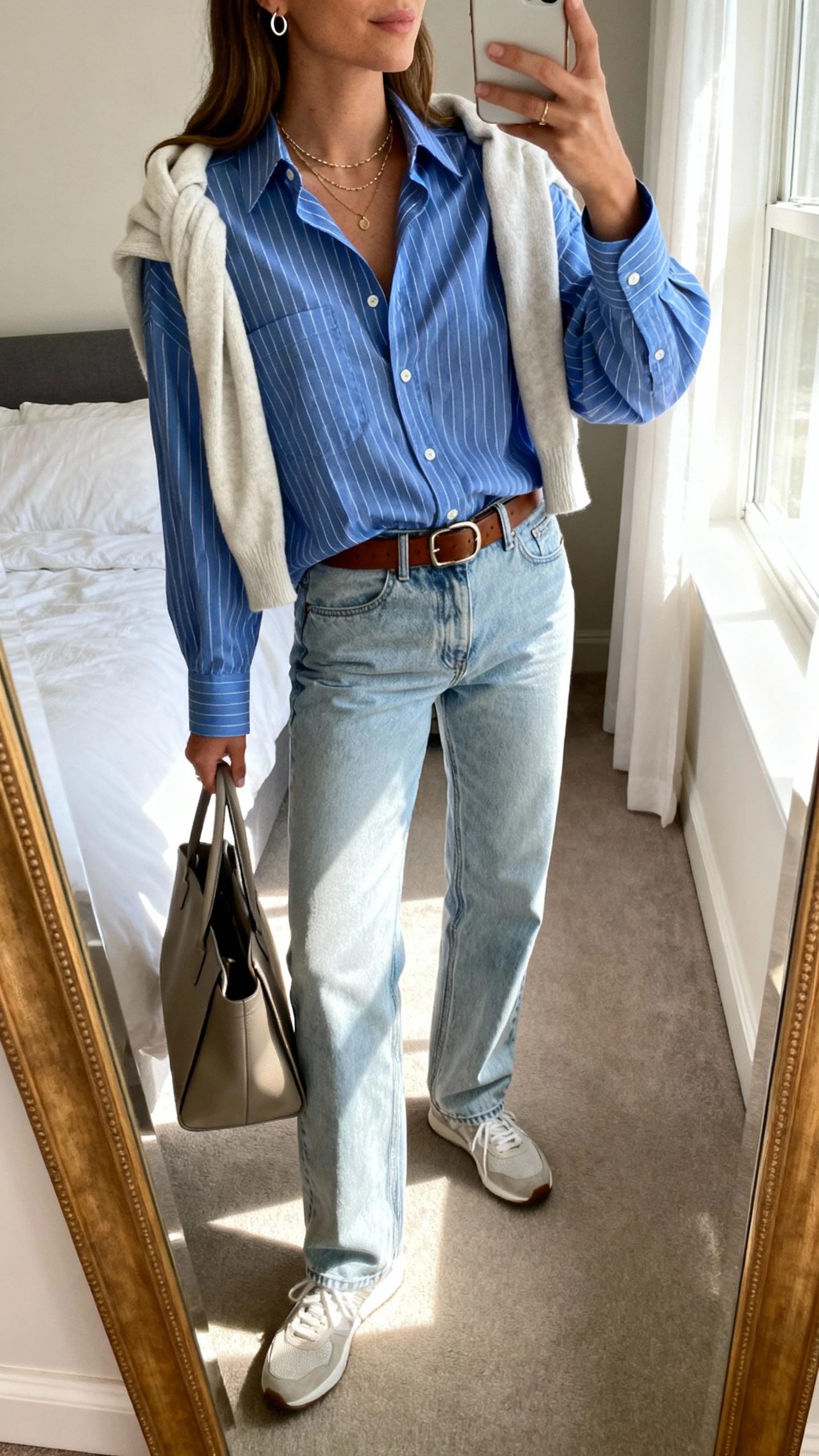 iPhone side-angle mirror selfie of a woman in an oversized blue striped button-down, light-wash straight-leg jeans, classic leather belt, minimal sneakers, layered delicate necklaces, small hoops, and a light sweater tied over the shoulders, holding a structured tote; face not visible, bedroom mirror, morning window light, iPhone photo quality.