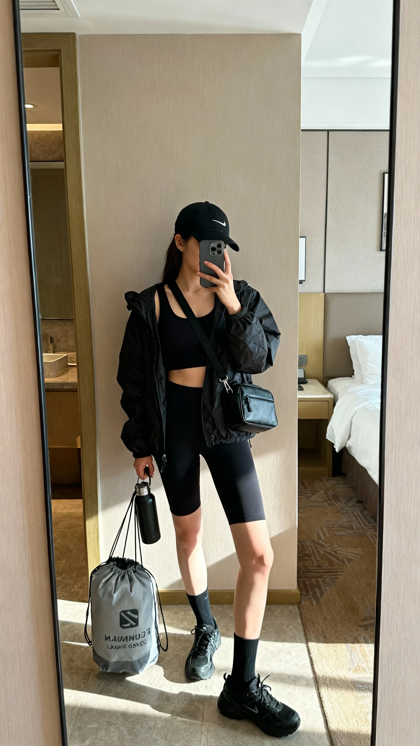 iPhone side-angle mirror selfie of a woman packing a black sports bra with black tank, black 7/8 leggings and black biker shorts, black lightweight packable jacket, black all-purpose trainers, black crew socks and ankle socks, black cap, compact black crossbody bag, travel-sized laundry bag, and a folding water bottle, face not visible, hotel room full-length mirror, morning light, iPhone photo quality.