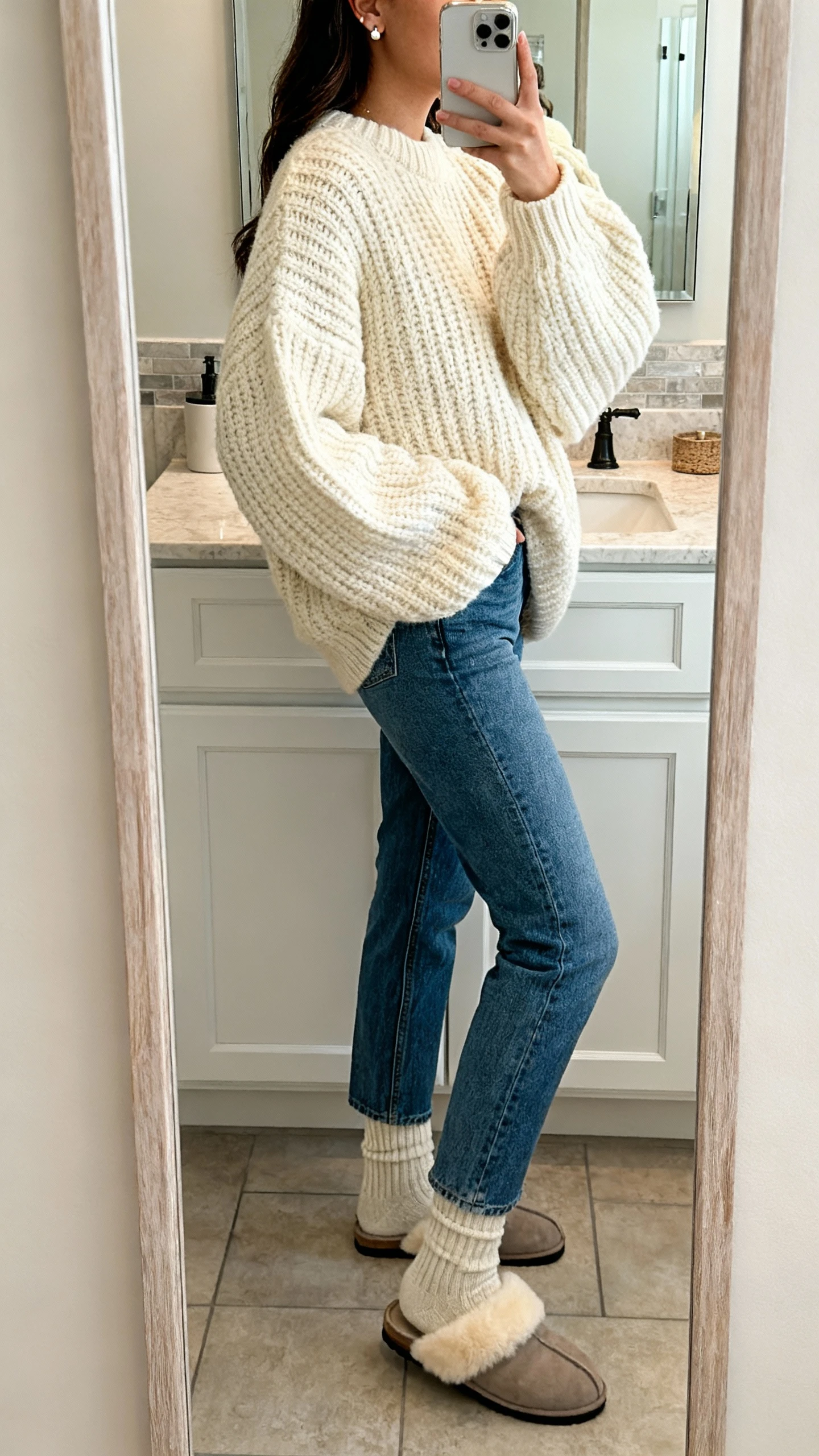 iPhone side-angle mirror selfie of a woman wearing mom jeans, oversized chunky knit sweater in cream, cozy wool socks, shearling-lined slippers, delicate stud earrings, face not visible, bathroom mirror, soft indoor lighting, iPhone photo quality