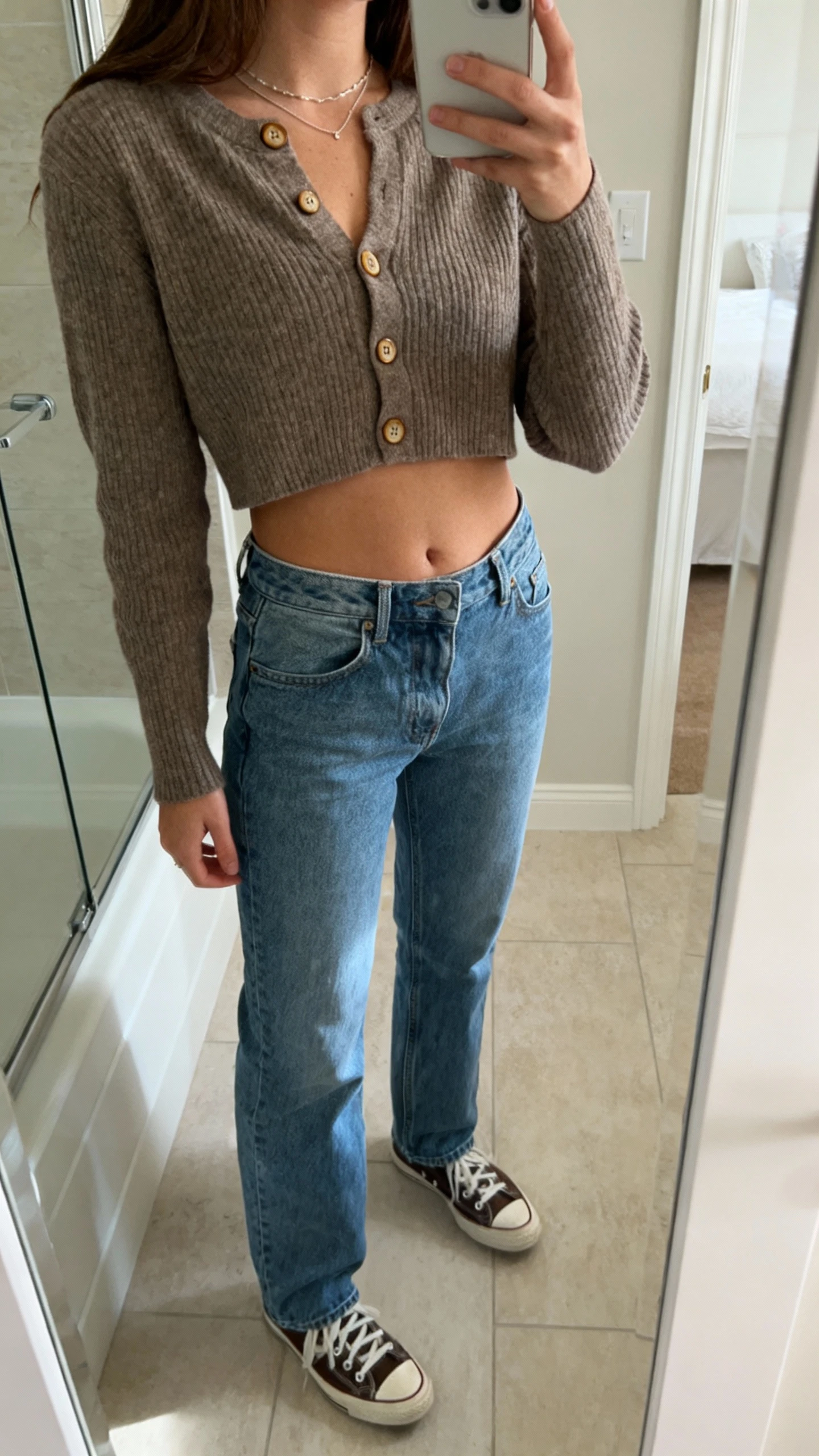 iPhone side-angle mirror selfie of a woman wearing mom jeans, a cropped cardigan buttoned at the center, delicate necklace, and retro sneakers, face not visible, cropped at shoulders, bathroom mirror, soft morning light, iPhone photo quality.