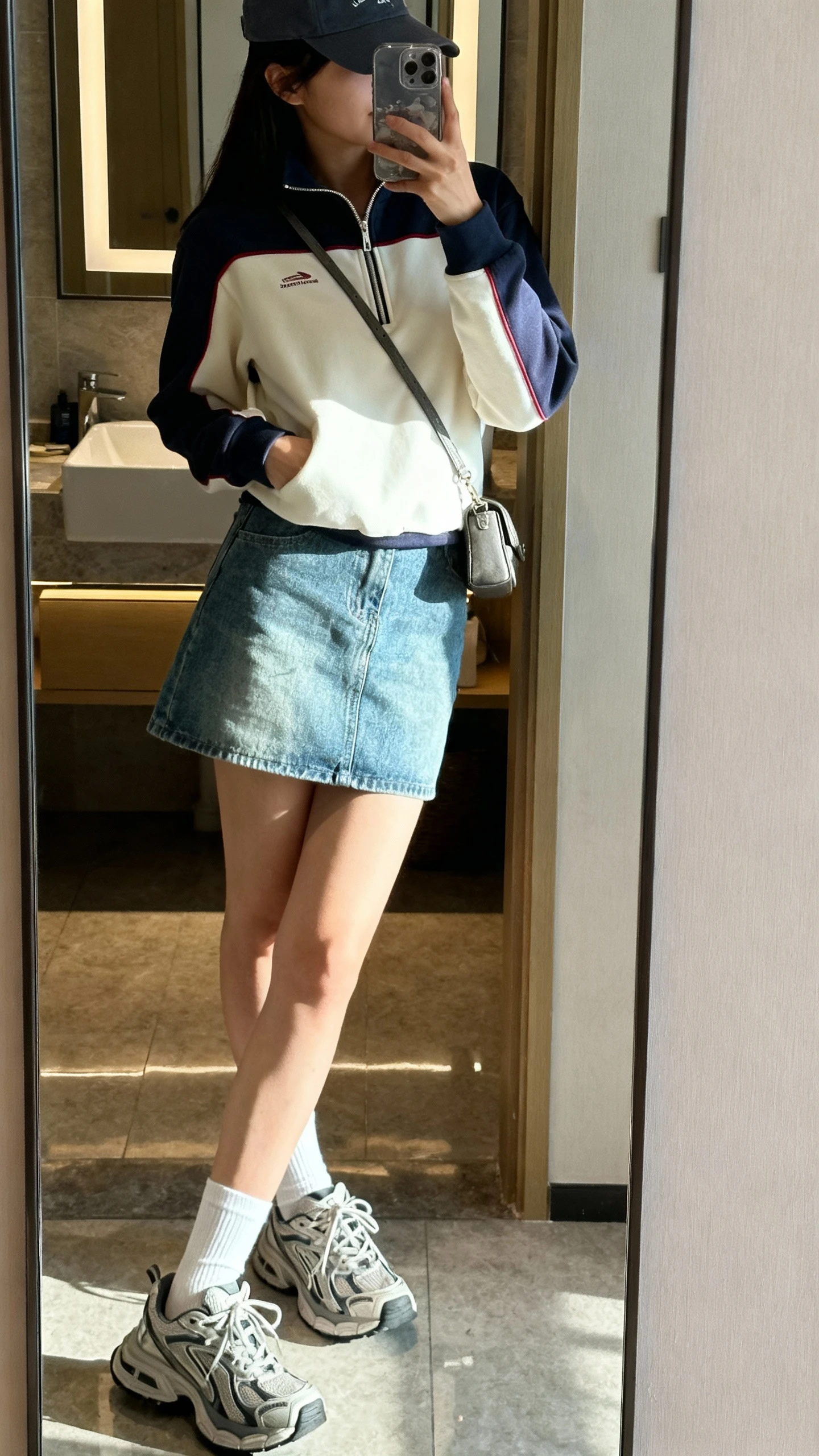 iPhone side-angle mirror selfie of a woman wearing a sporty half-zip pullover, denim mini skirt, white crew socks, chunky athletic sneakers, baseball cap, and a small crossbody, face not visible, bathroom mirror with indoor morning light, iPhone photo quality.