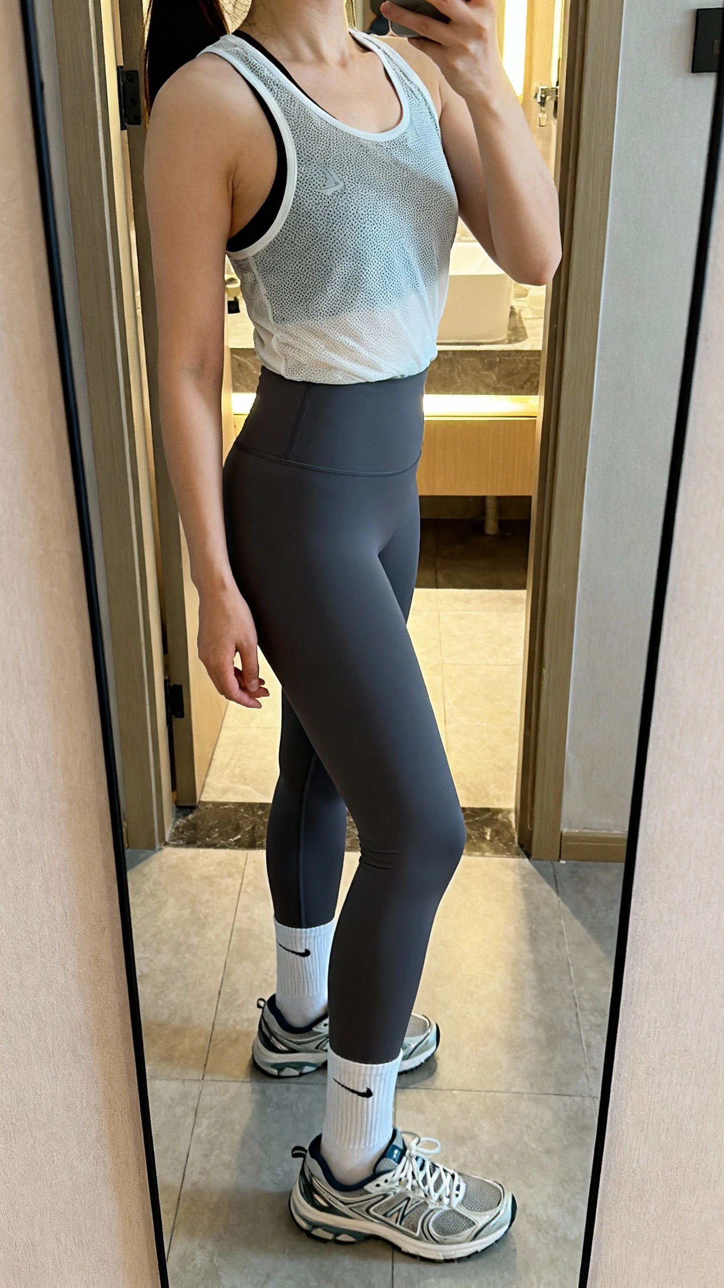 iPhone side-angle mirror selfie of a woman wearing a lightweight sweat-wicking mesh tank, quick-dry high-rise leggings, and breathable running socks with trainers, face not visible, cropped at shoulders, bathroom mirror with indoor lighting, iPhone photo quality