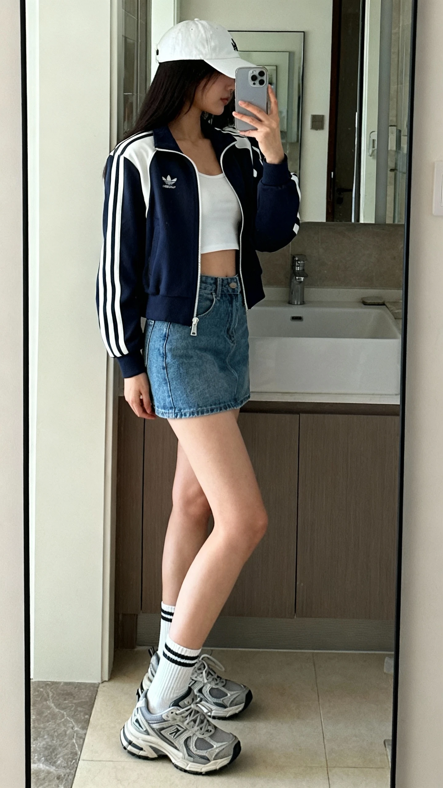 iPhone side-angle mirror selfie of a woman wearing a denim mini skirt, sporty zip-up track jacket, white crop top, baseball cap, crew socks, and chunky trainers, face not visible, bathroom mirror, indoor morning light, iPhone photo quality.