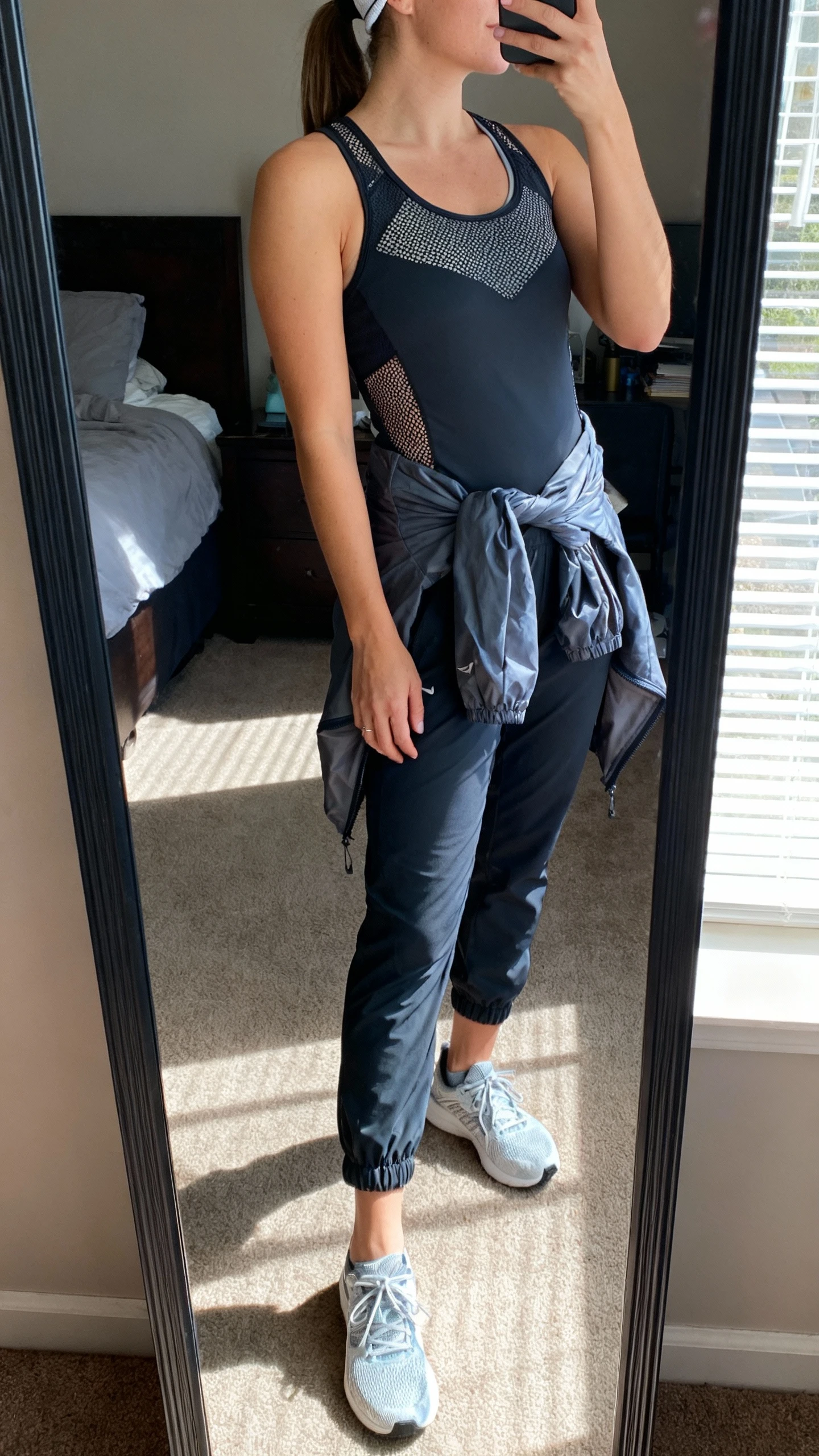iPhone side-angle mirror selfie of a woman wearing a fitted performance tank with mesh panels, tapered training joggers with ankle cuffs, lightweight cross-training shoes, a packable windbreaker tied around the waist, minimal headband, face not visible, bedroom full-length mirror with natural window light, iPhone photo quality