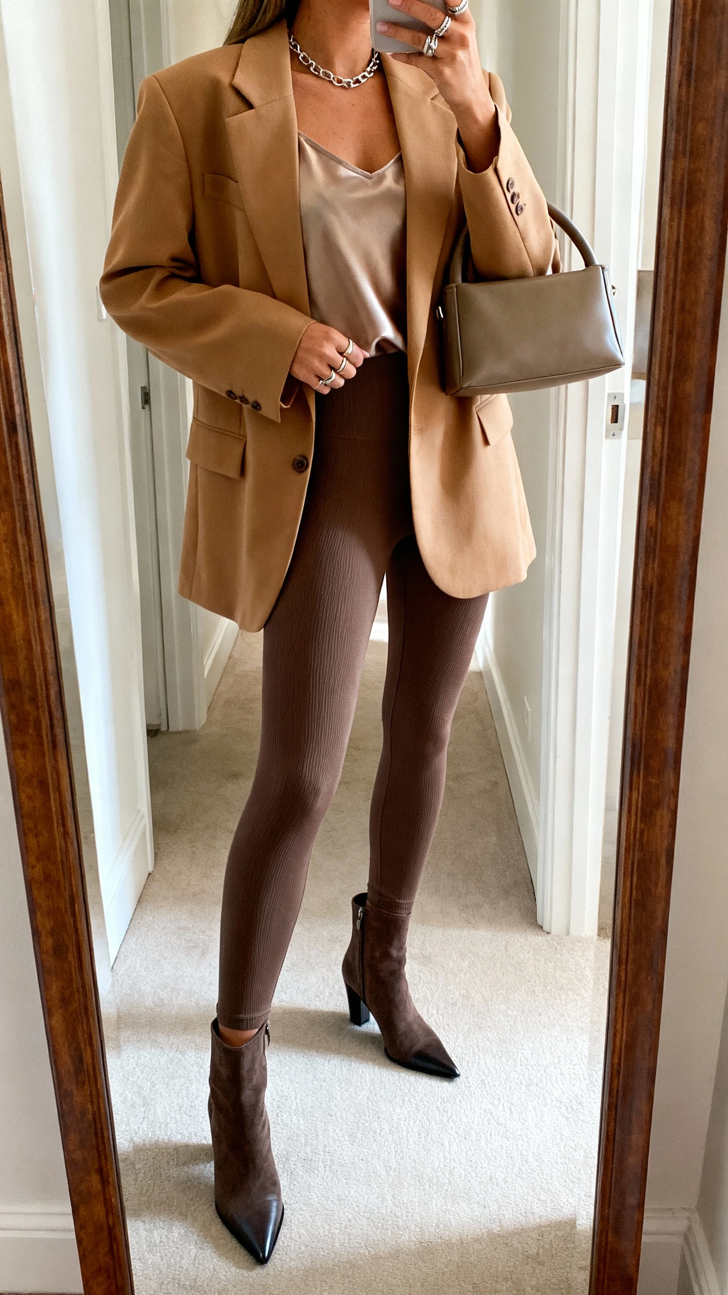 iPhone side-angle mirror selfie of a woman wearing an oversized camel blazer, thick ankle-length ponte leggings, pointed ankle boots, a silky tank, structured top-handle bag, delicate chain necklace and stacked rings, face not visible, cropped at shoulders, hallway full-length mirror, soft daylight, iPhone photo quality.