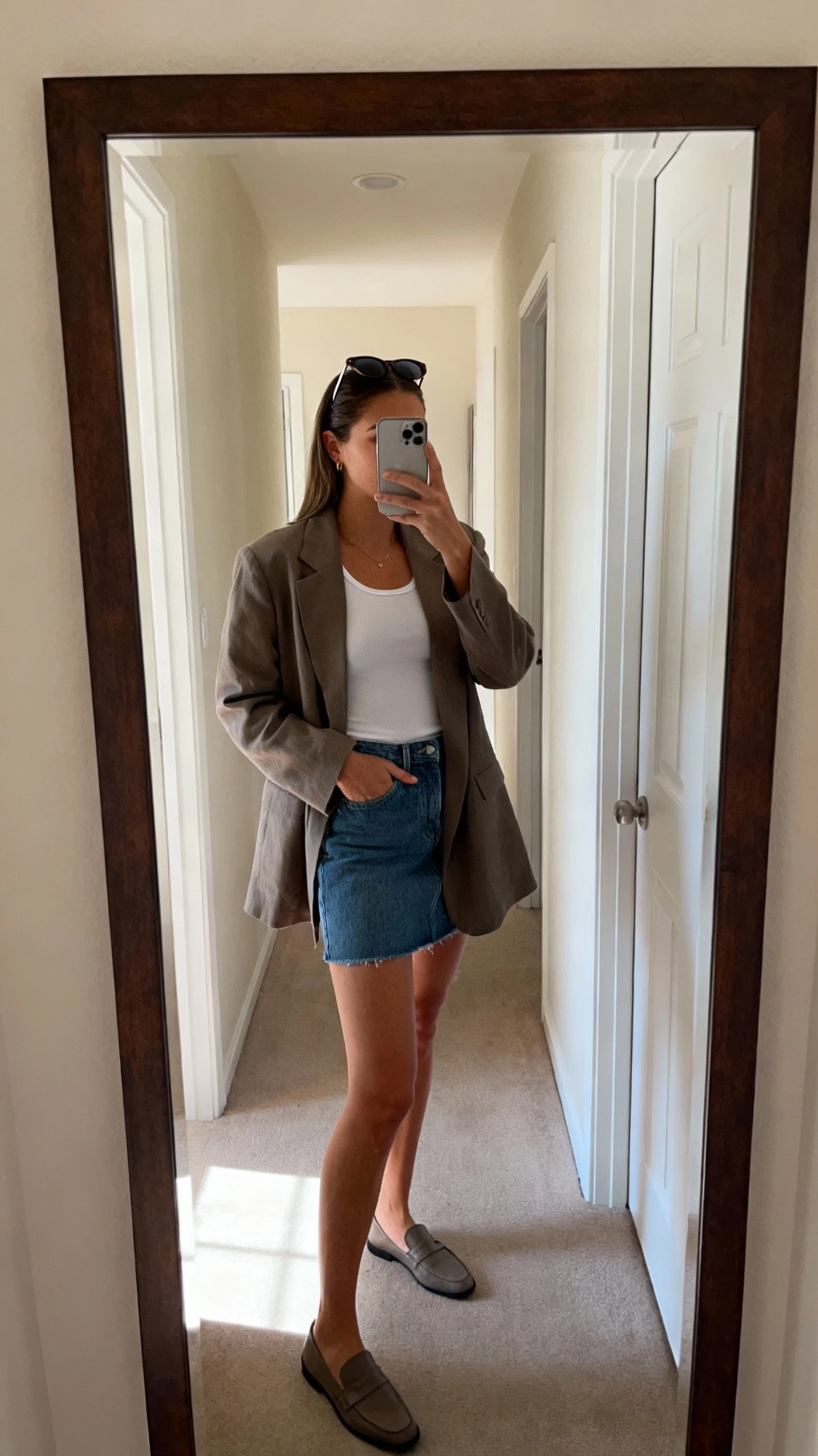 iPhone side-angle mirror selfie of a woman wearing a relaxed-fit blazer over a white fitted tank, denim mini skirt, loafers, and slim sunglasses perched on head, face not visible, full-length hallway mirror with natural daylight, iPhone photo quality.