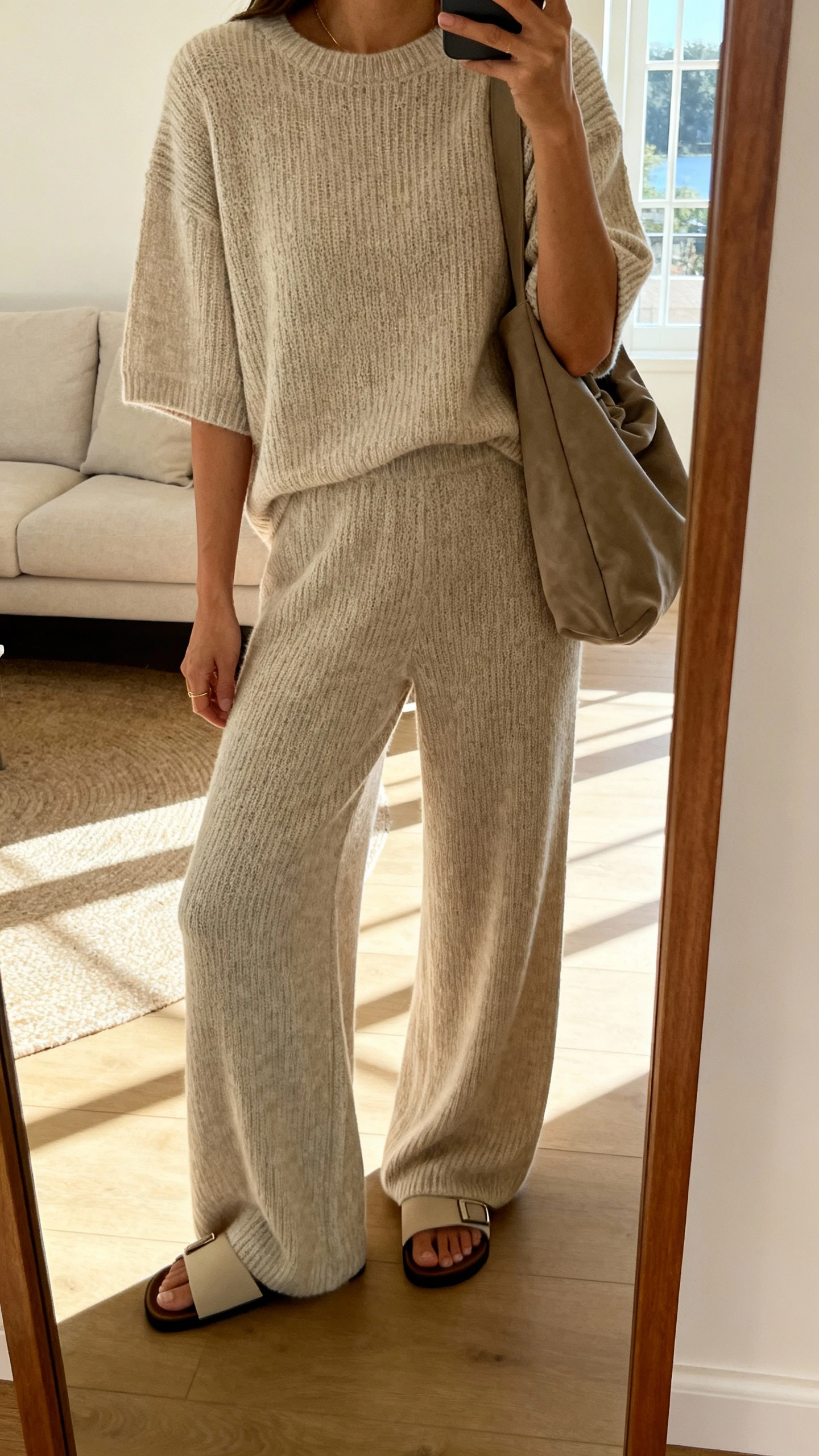 iPhone side-angle mirror selfie of a woman wearing a luxe matching knit set (boxy short-sleeve sweater and wide-leg knit pants), slide sandals, and a slouchy shoulder bag, face not visible, cropped at shoulders, living room mirror with natural light, iPhone photo quality.