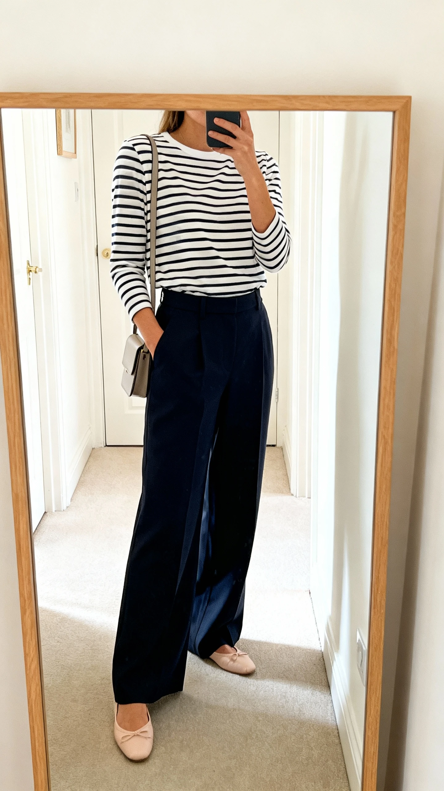 iPhone side-angle mirror selfie of a woman wearing a Breton striped long-sleeve tee tucked into high-waisted wide-leg trousers, ballet flats, and a slim shoulder bag, face not visible, cropped at shoulders, hallway mirror, bright indoor daylight, iPhone photo quality.