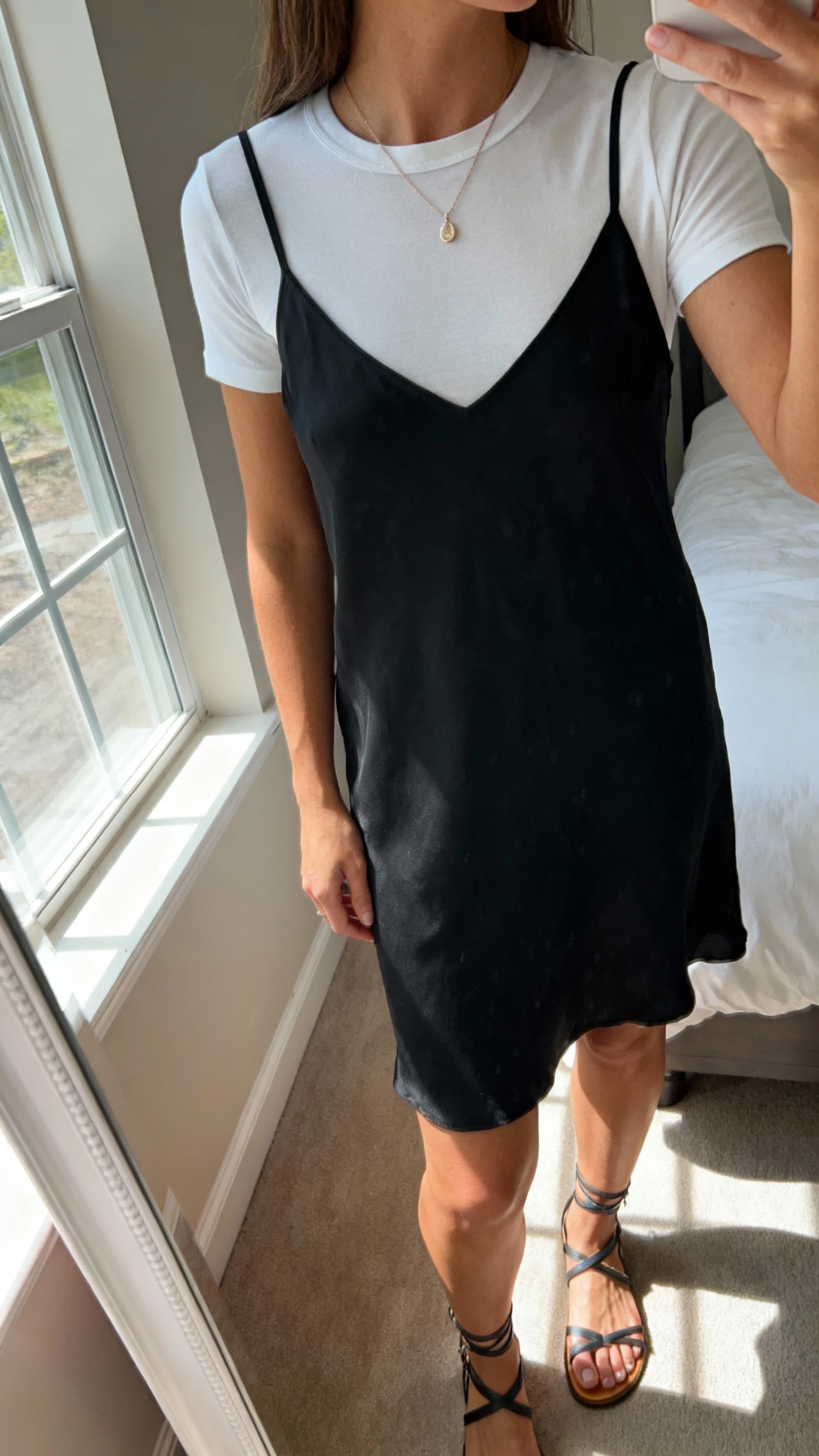 iPhone side-angle mirror selfie of a woman wearing a black slip dress layered over a fitted white tee, delicate pendant necklace, strappy sandals, face not visible, cropped at shoulders, bedroom mirror, natural window light, iPhone photo quality