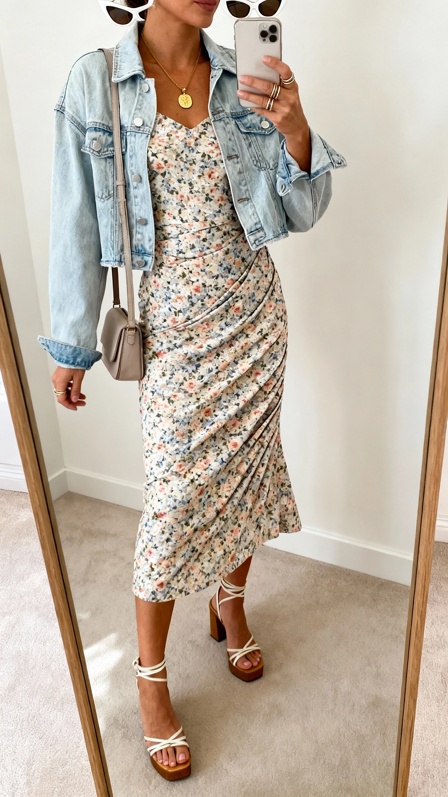 iPhone side-angle mirror selfie of a woman wearing a floral bias-cut midi dress, cropped light-wash denim jacket, strappy block-heel sandals, small shoulder bag, gold pendant necklace, stackable rings, cat-eye sunglasses on head; face not visible, cropped at neck, full-length mirror, indoor daylight, iPhone photo quality.