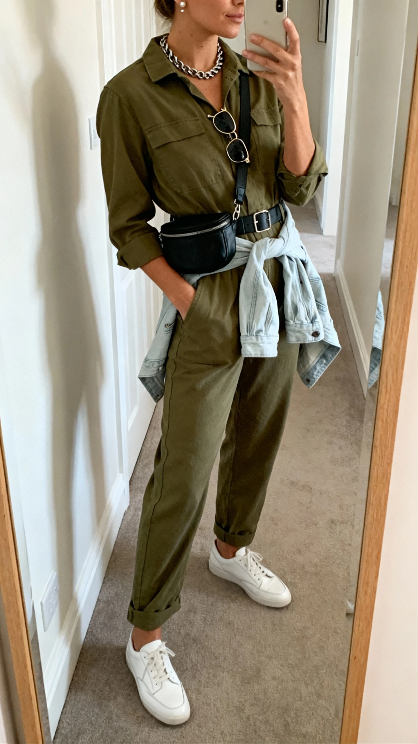 iPhone side-angle mirror selfie of a woman wearing an olive relaxed-fit utility jumpsuit, clean white leather sneakers, black belt bag crossbody, chunky chain necklace, small stud earrings, classic aviator sunglasses tucked on neckline, and a light denim jacket tied at the waist, face not visible, hallway full-length mirror, natural daylight, iPhone photo quality.