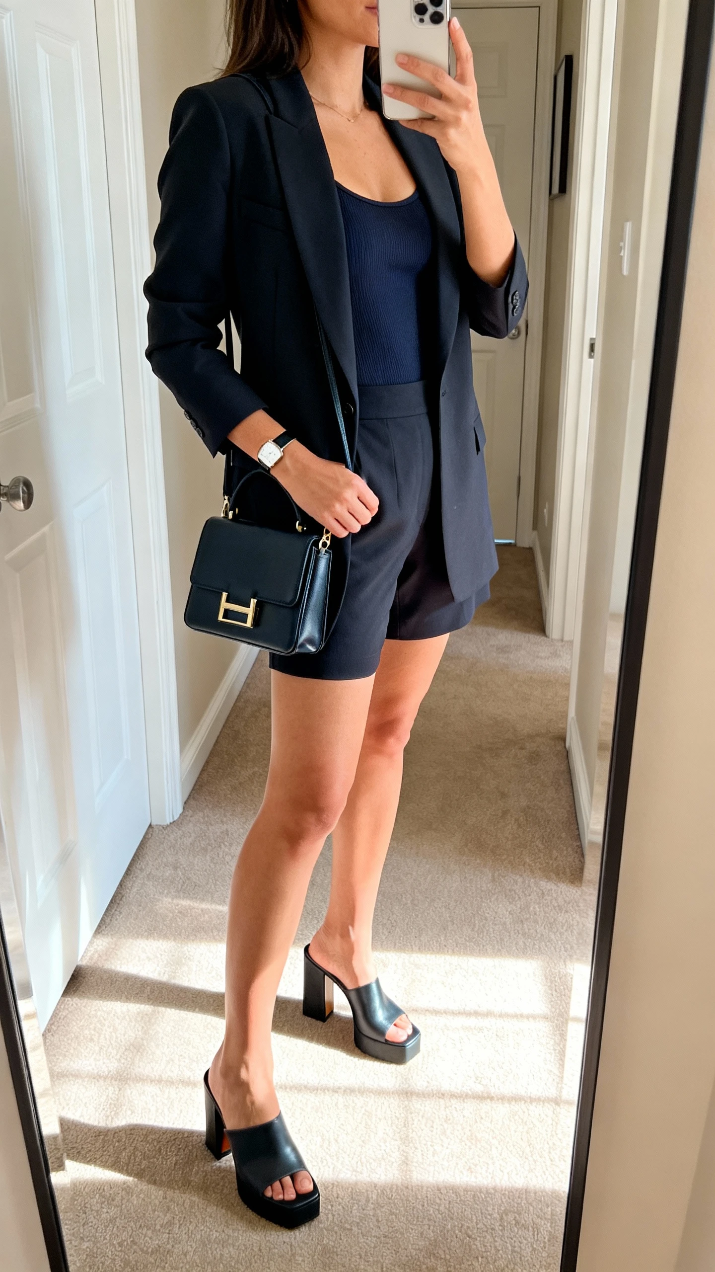 iPhone side-angle mirror selfie of a woman wearing a tailored shorts suit (matching blazer and tailored shorts) with a fitted camisole, block-heel mules, structured mini tote, slim watch, face not visible, hallway mirror, indoor daylight, iPhone photo quality.