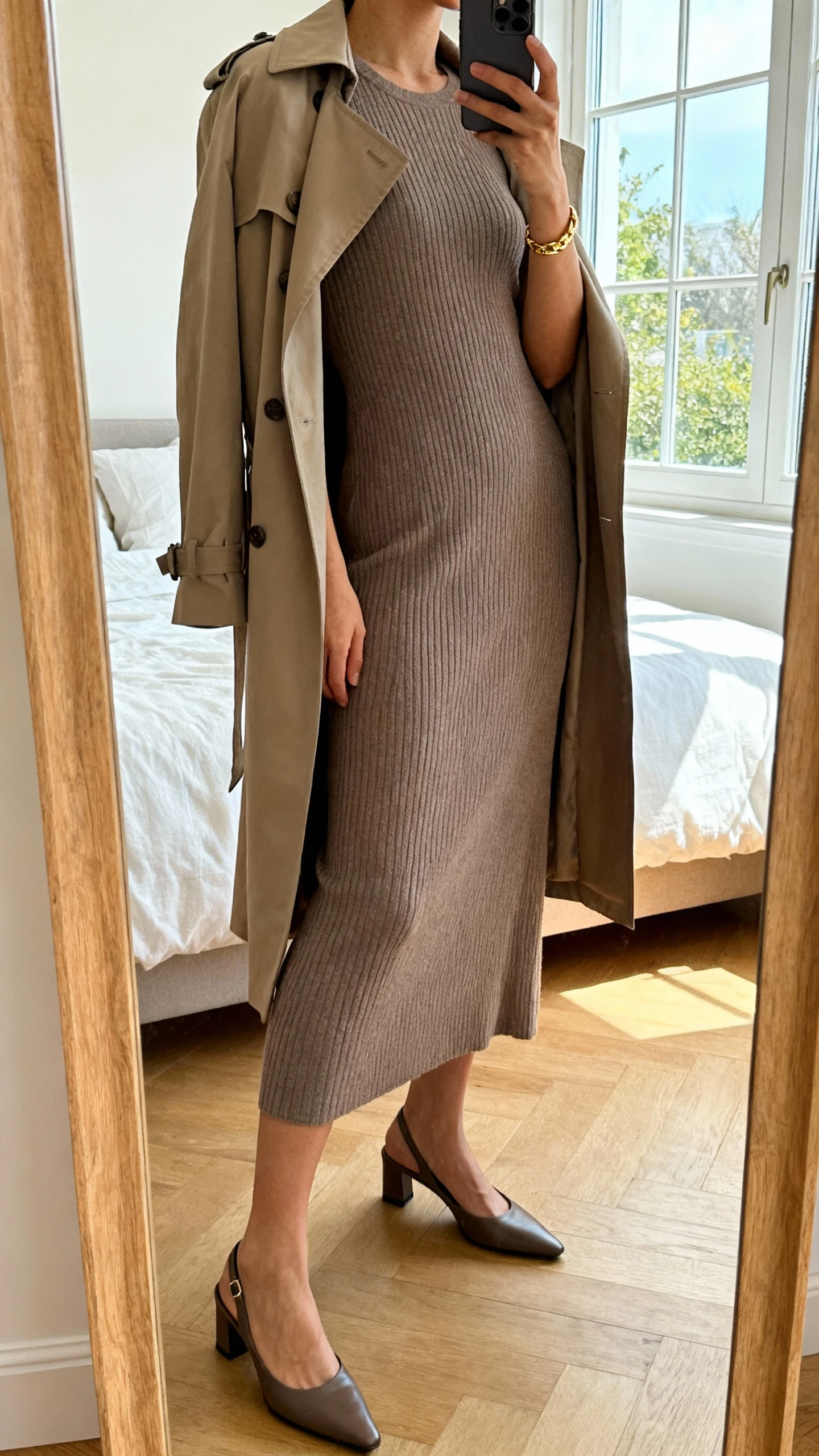 iPhone side-angle mirror selfie of a woman wearing a soft ribbed knit midi dress in taupe with a classic trench draped over shoulders, sleek slingback flats, minimal gold bracelet, face not visible, cropped at neck, bedroom mirror, natural window light, iPhone photo quality.