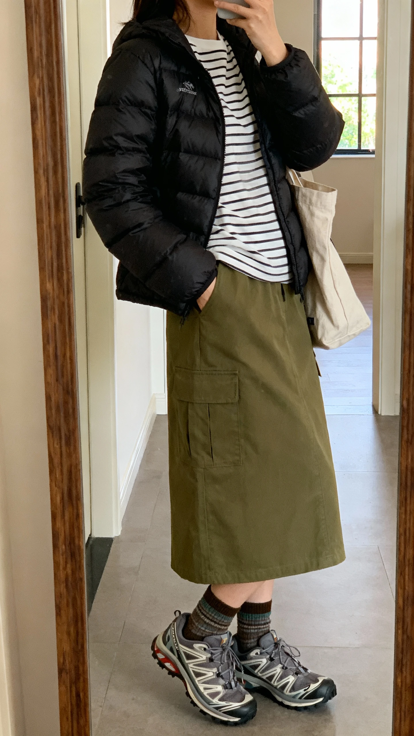 iPhone side-angle mirror selfie of a woman wearing an olive utility midi skirt with pockets, lightweight black puffer jacket over a striped tee, trail-inspired sneakers, canvas tote, crew socks, face not visible, entryway mirror, indoor window light, iPhone photo quality.