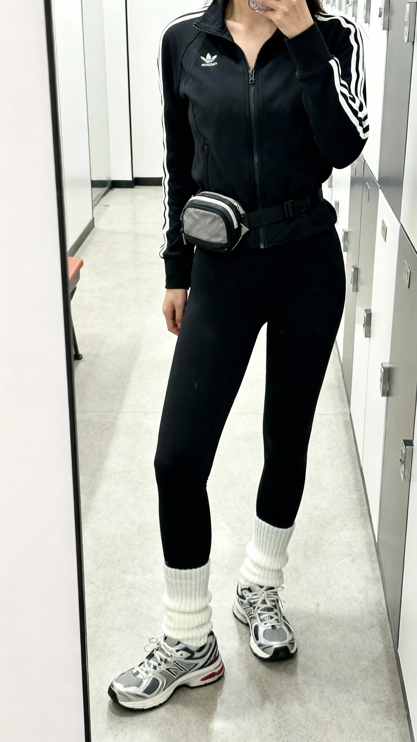 iPhone side-angle mirror selfie of a woman wearing black performance leggings, a sporty zip-up track jacket, crisp white crew socks pulled up, retro running sneakers, and a small belt bag, face not visible, cropped at shoulders, gym locker-room mirror, bright indoor light, iPhone photo quality.