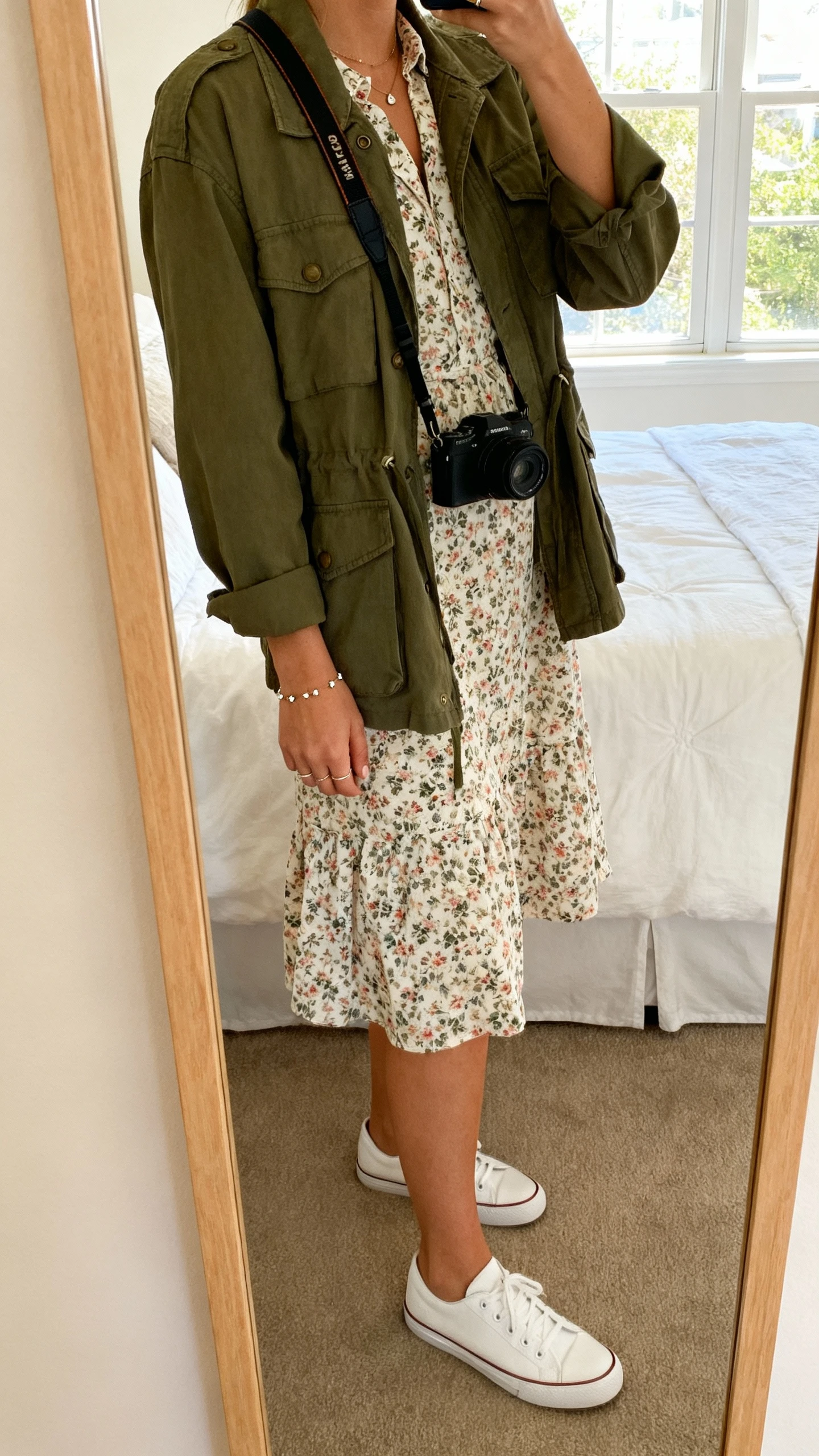 iPhone side-angle mirror selfie of a woman wearing a floral day dress layered with an olive utility jacket, crossbody camera bag, dainty studs, white low-top sneakers, face not visible, bedroom mirror, natural window light, iPhone photo quality.
