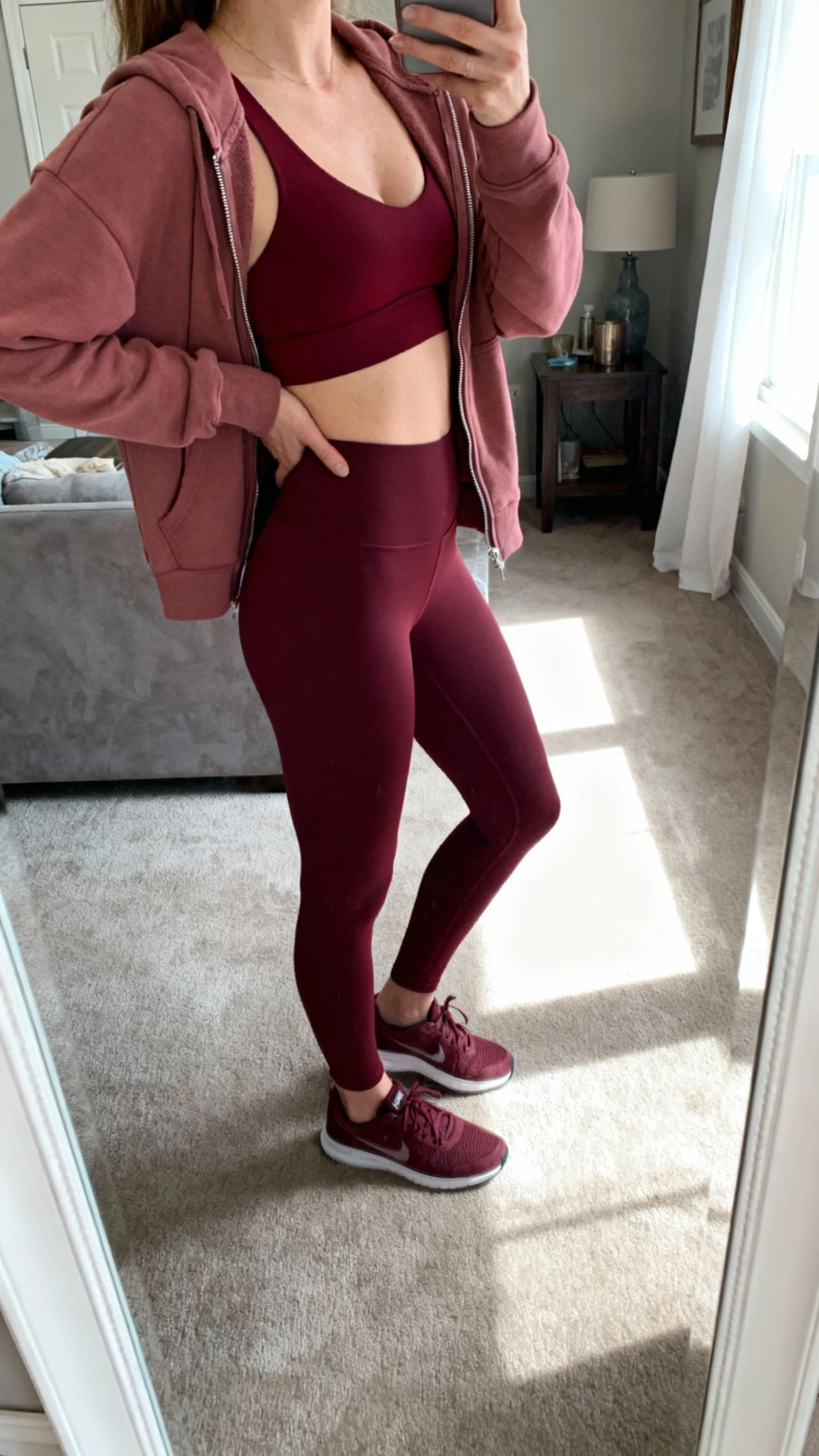 iPhone side-angle mirror selfie of a woman wearing burgundy leggings, a burgundy longline bra, dusty pink zip hoodie, and burgundy sneakers, face not visible, living room mirror, soft morning light, iPhone photo quality.