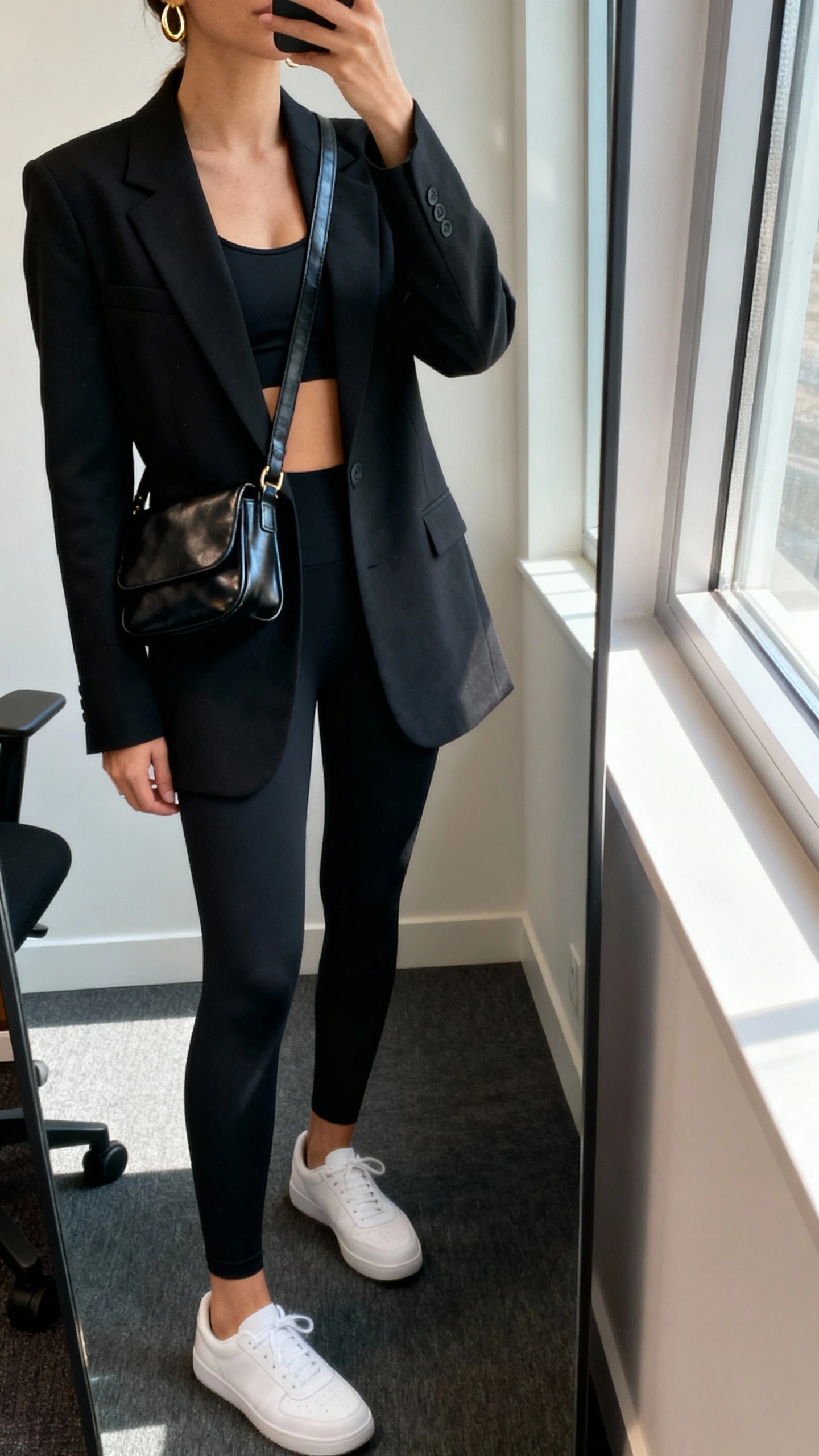 iPhone side-angle mirror selfie of a woman wearing a tailored black blazer over a black sports bra and leggings, sleek leather crossbody, minimalist white sneakers, and subtle gold hoops, face not visible, cropped at neck, office corner mirror, window light, iPhone photo quality.
