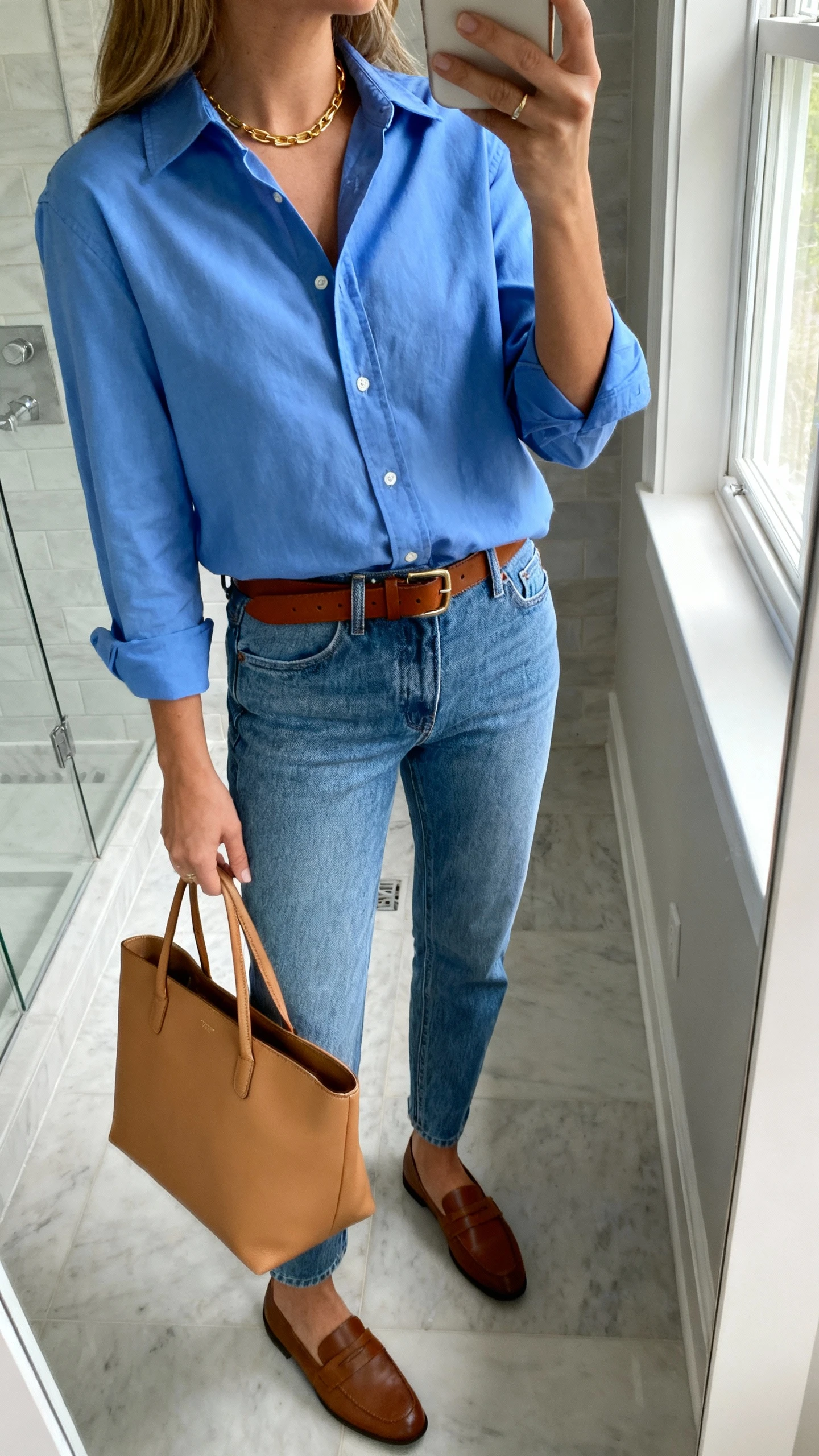 iPhone side-angle mirror selfie of a woman wearing mom jeans, crisp blue button-up shirt tucked in, brown leather belt, loafers, structured tan tote, simple gold necklace, face not visible, bathroom mirror, natural window light, iPhone photo quality