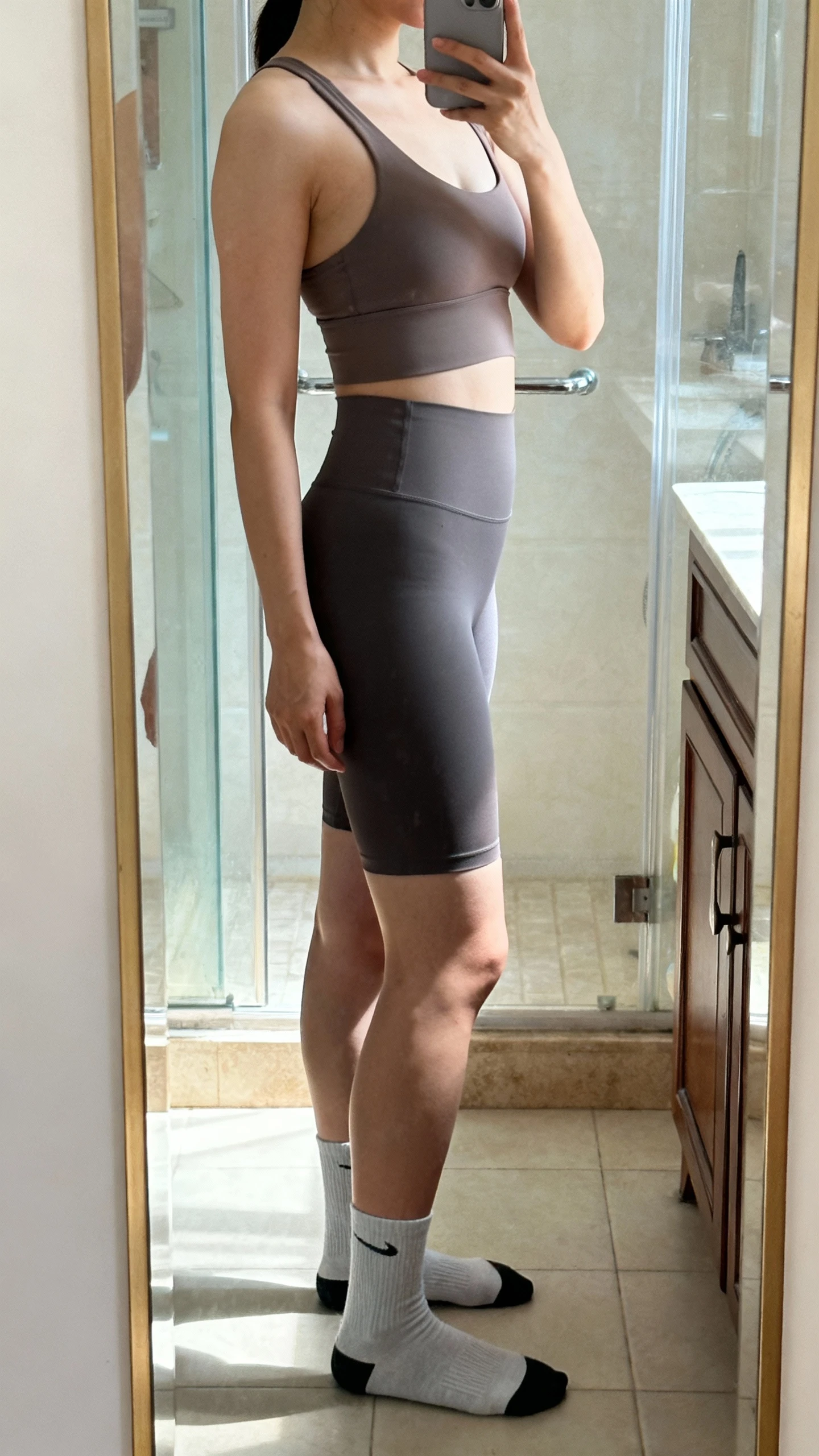 iPhone side-angle mirror selfie of a woman wearing seamless anti-chafe biker shorts, a seamless longline bra, and no-show performance socks, face not visible, cropped at shoulders, bathroom mirror, natural daylight, iPhone photo quality