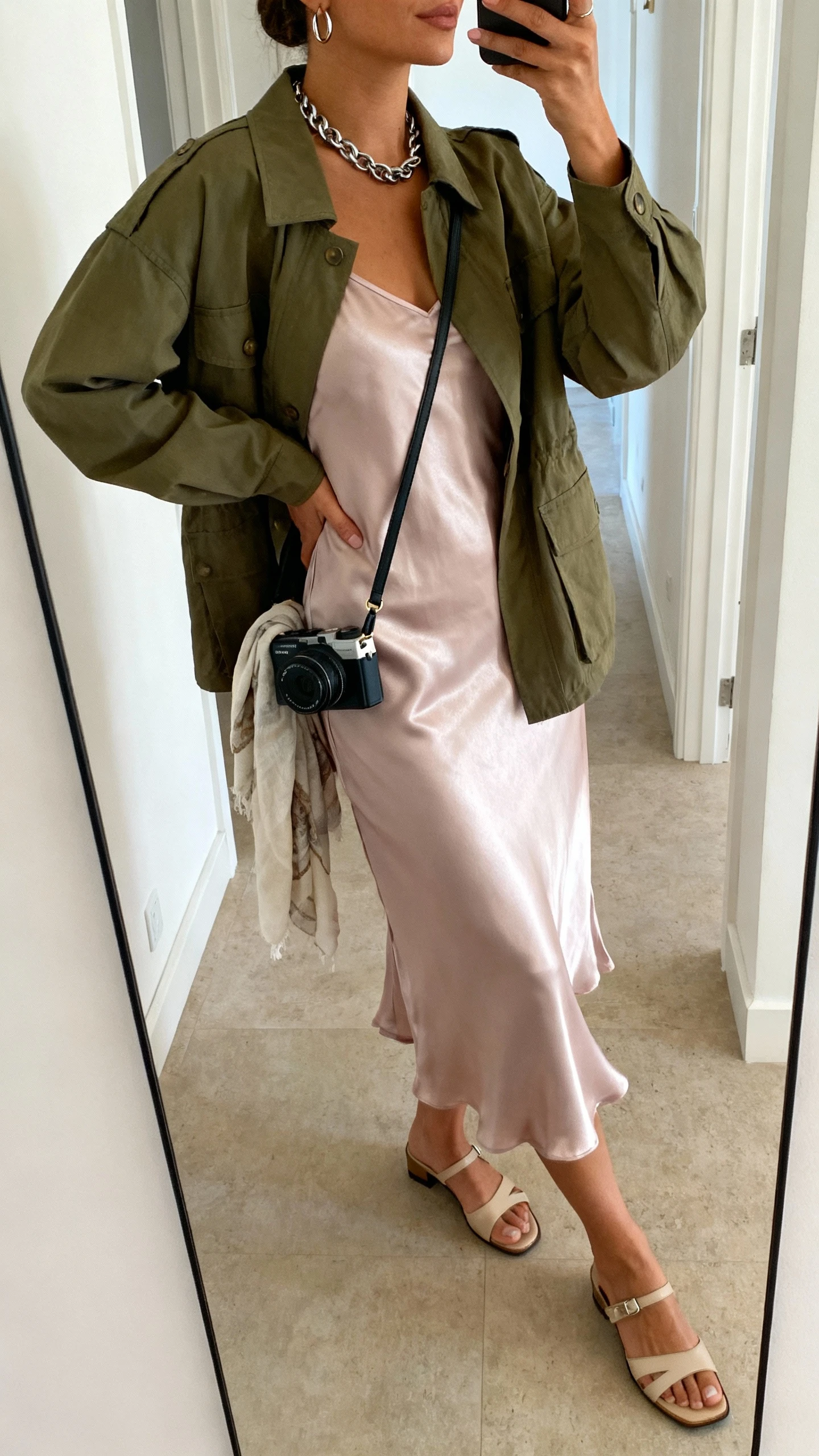 iPhone side-angle mirror selfie of a woman wearing a satin midi slip dress in pastel, olive utility jacket layered on top, low-heeled sandals, mini camera bag, chunky chain necklace, small hoops, lightweight scarf draped over bag strap; face not visible, cropped at neck, hallway mirror, natural daylight, iPhone photo quality.