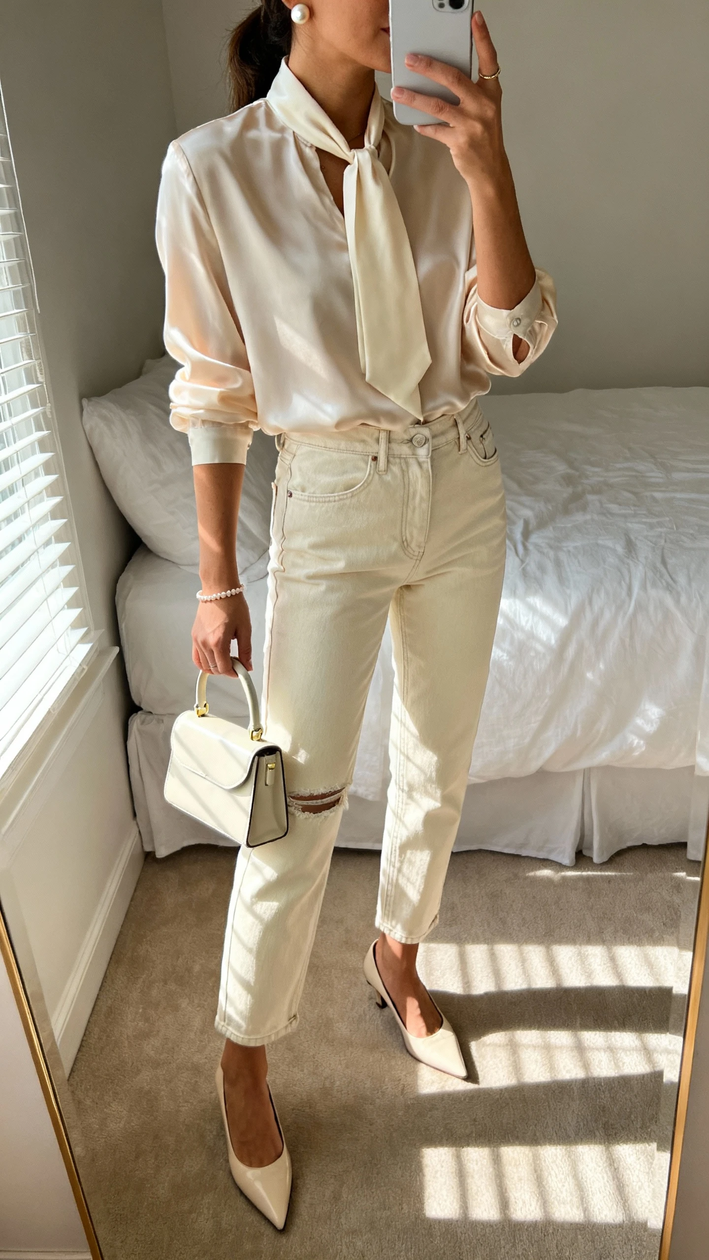 iPhone side-angle mirror selfie of a woman wearing a silk tie-neck blouse, ecru mom jeans (no distressing), pointed-toe heels, pearl studs, a delicate bracelet, and a mini top-handle bag, face not visible, cropped at neck, bedroom mirror with morning light, iPhone photo quality.