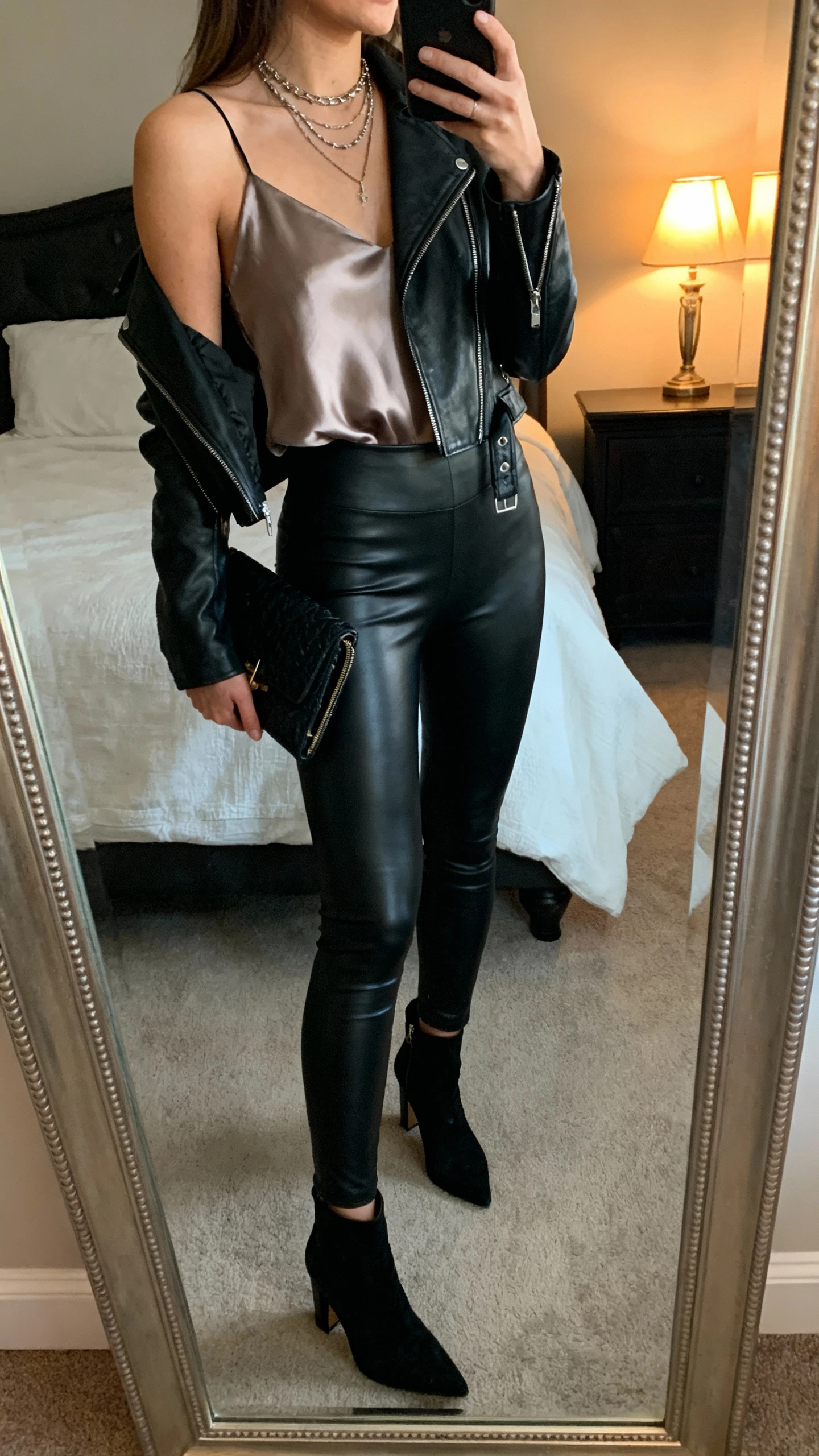 iPhone side-angle mirror selfie of a woman wearing leather-look black leggings, silky camisole, cropped moto jacket, heeled ankle boots, clutch, layered necklaces, face not visible, bedroom mirror with evening lamp light, iPhone photo quality