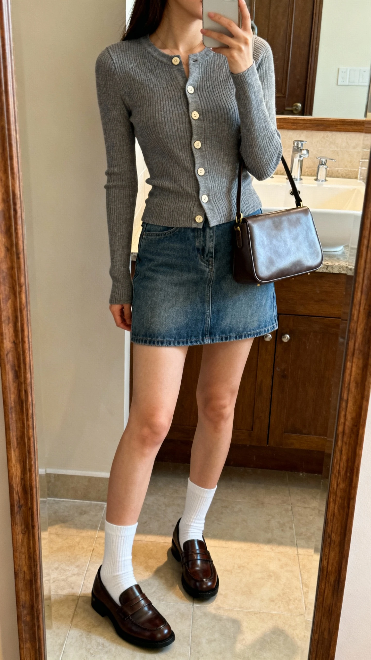 iPhone side-angle mirror selfie of a woman wearing a fitted cardigan buttoned like a top, denim mini skirt, polished loafers, white socks, and a structured satchel, face not visible, bathroom mirror with soft indoor light, iPhone photo quality.