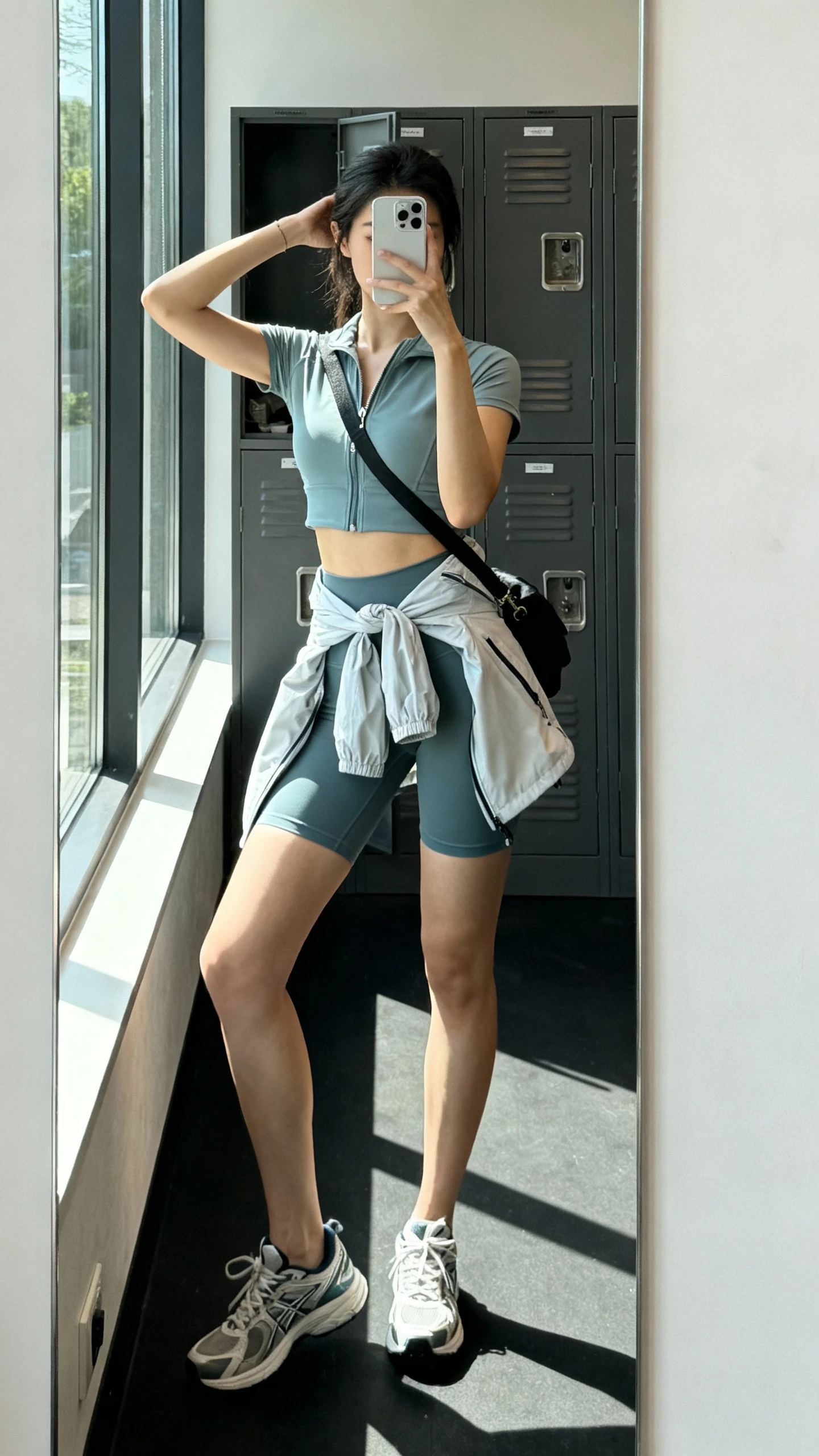 iPhone side-angle mirror selfie of a woman wearing an athletic-chic matching set (high-waist biker shorts and zip-up or cropped sports top), lightweight windbreaker tied at waist, running sneakers, sporty crossbody, face not visible, gym locker mirror, natural daylight from window, iPhone photo quality.