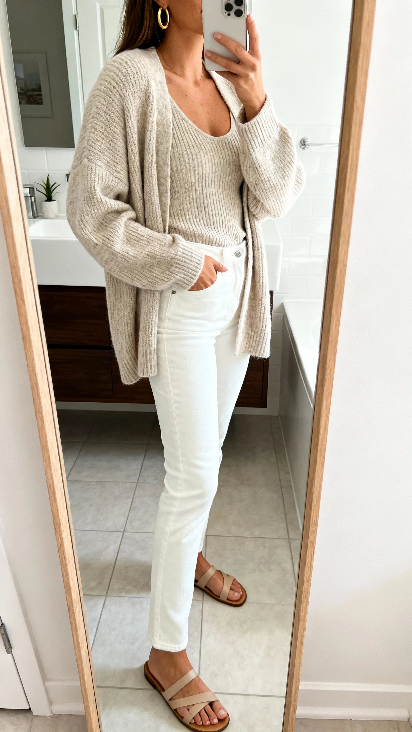 iPhone side-angle mirror selfie of a woman wearing tonal knit sweater layered over a matching knit tank, white straight jeans, minimal sandals, and gold hoops, face not visible, bathroom full-length mirror, bright indoor light, iPhone photo quality.