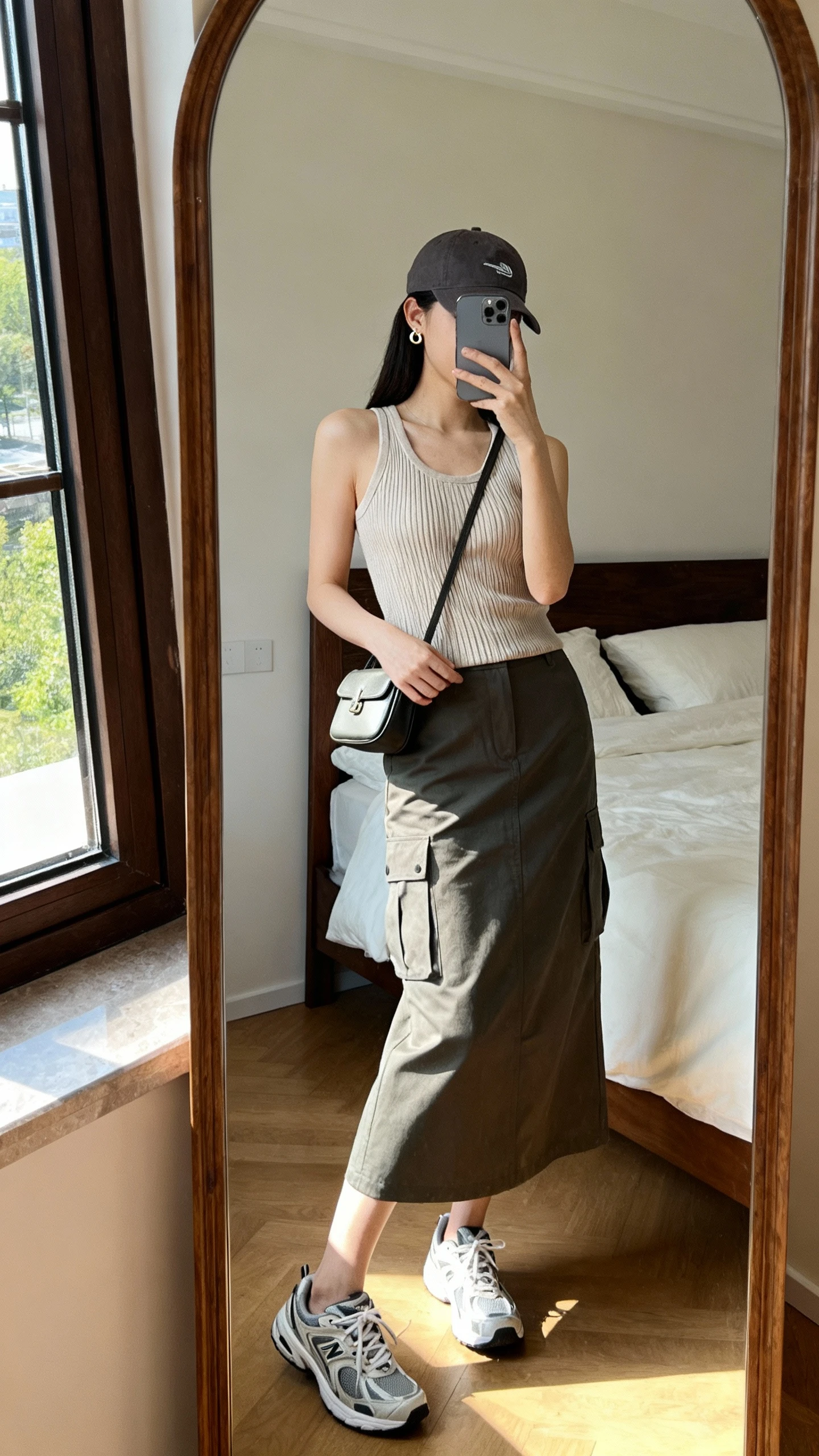 iPhone side-angle mirror selfie of a woman wearing a ribbed knit tank with a midi cargo skirt, sporty dad sneakers, baseball cap, tiny huggie earrings, and a mini shoulder bag, face not visible, bedroom full-length mirror, afternoon window light, iPhone photo quality