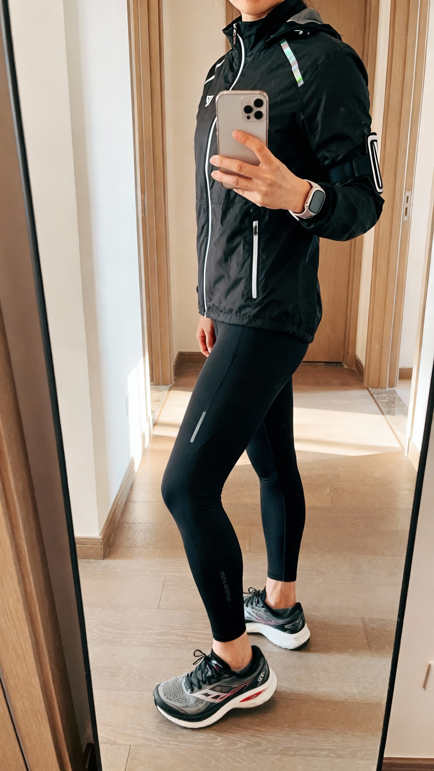 iPhone side-angle mirror selfie of a woman wearing performance black running leggings and a race-ready black shell jacket with reflective accents, face not visible, hallway mirror, morning light, running shoes and slim armband, iPhone photo quality.