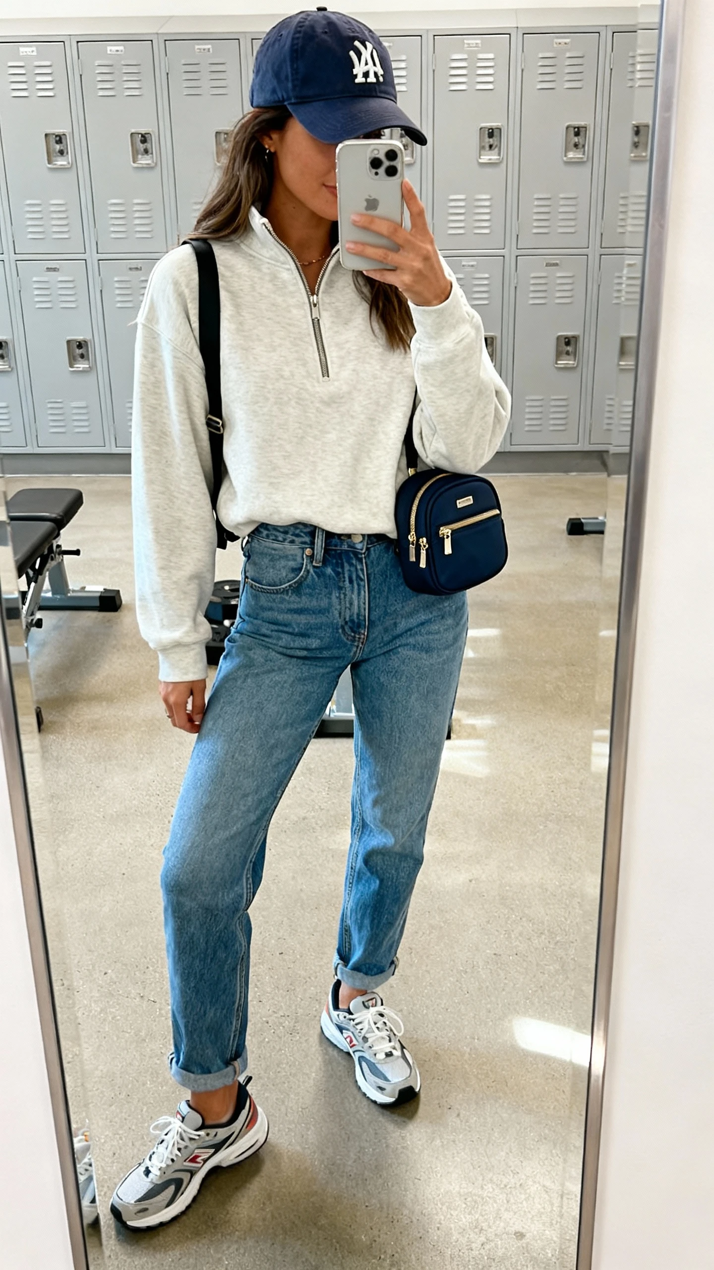 iPhone side-angle mirror selfie of a woman wearing mom jeans, a half-zip pullover, a baseball cap, sporty sneakers, and a mini backpack, face not visible, cropped at shoulders, gym locker mirror, bright indoor lighting, iPhone photo quality.