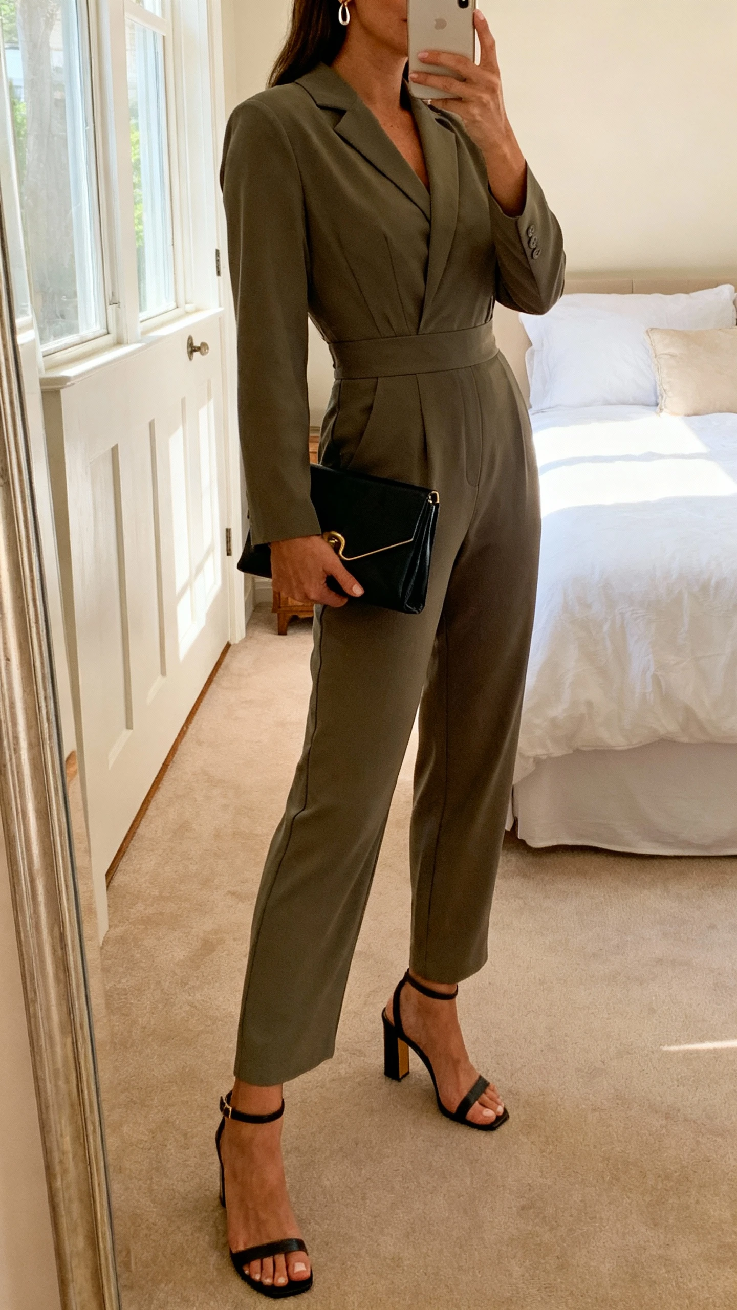 iPhone side-angle mirror selfie of a woman wearing an elevated tailored jumpsuit with a defined waist, heeled sandals, structured clutch, subtle earrings, face not visible, bedroom mirror, warm indoor lighting, iPhone photo quality.