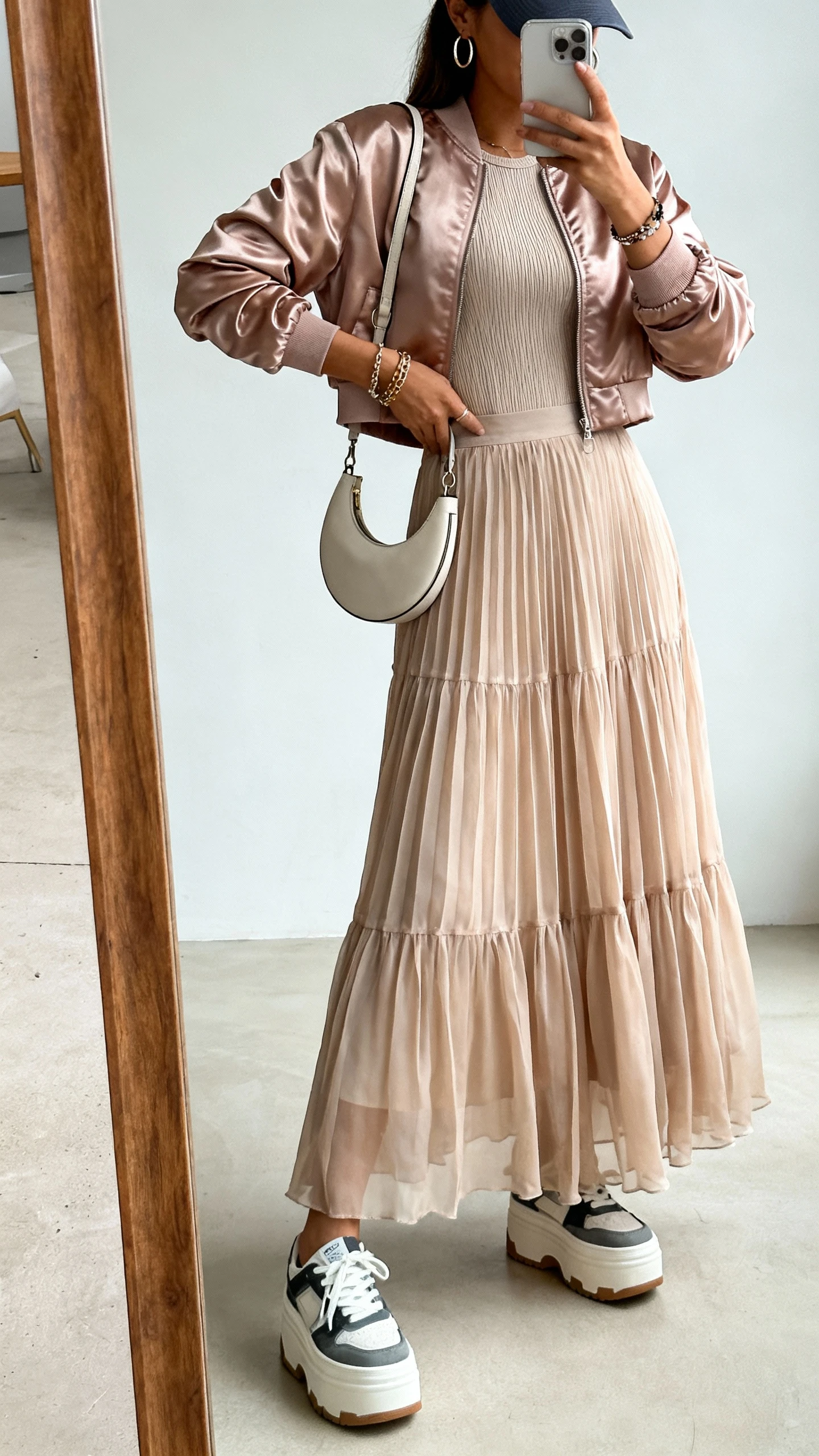 iPhone side-angle mirror selfie of a woman wearing a flowy chiffon maxi skirt, satin cropped bomber jacket over a fitted ribbed top, platform sneakers, crescent shoulder bag, huggie hoops, layered bracelets, and a baseball cap; face not visible, cropped at shoulders, full-length mirror, natural daylight, iPhone photo quality.