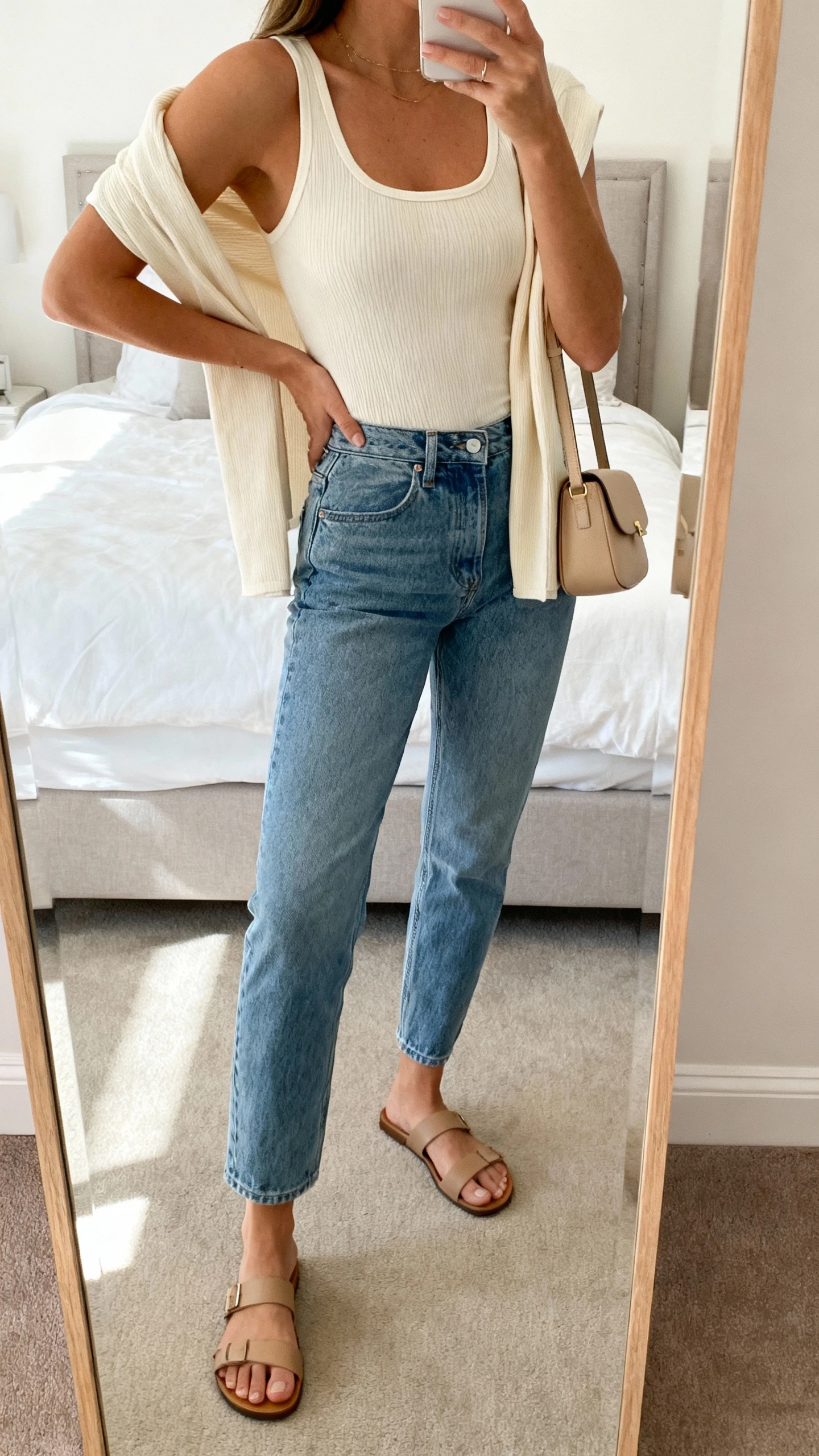iPhone side-angle mirror selfie of a woman wearing mom jeans, fitted ribbed tank in cream, matching lightweight cardigan draped over shoulders, minimalist leather sandals, small beige shoulder bag, face not visible, cropped at shoulders, bedroom mirror, soft morning light, iPhone photo quality