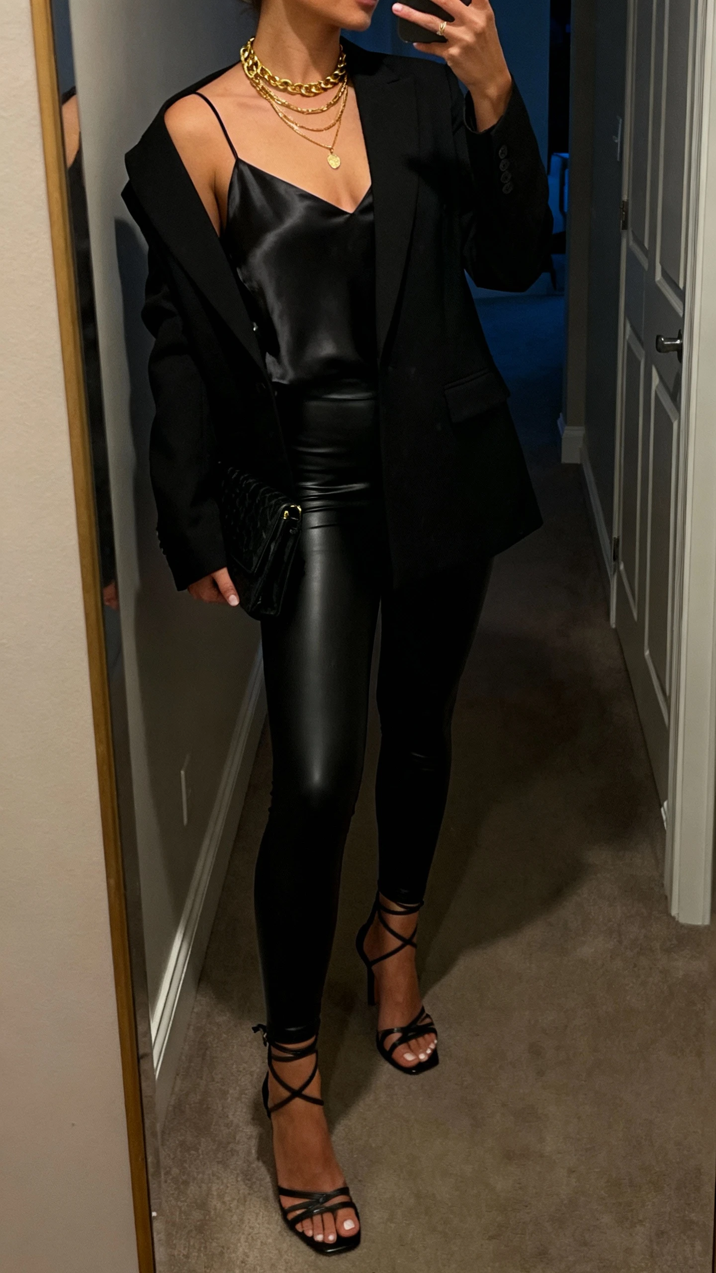 iPhone side-angle mirror selfie of a woman wearing faux-leather leggings after-dark: black faux-leather leggings, satin camisole, sharp black blazer, strappy heeled sandals, clutch, layered gold necklaces, face not visible, hallway full-length mirror, dim evening indoor lighting, iPhone photo quality