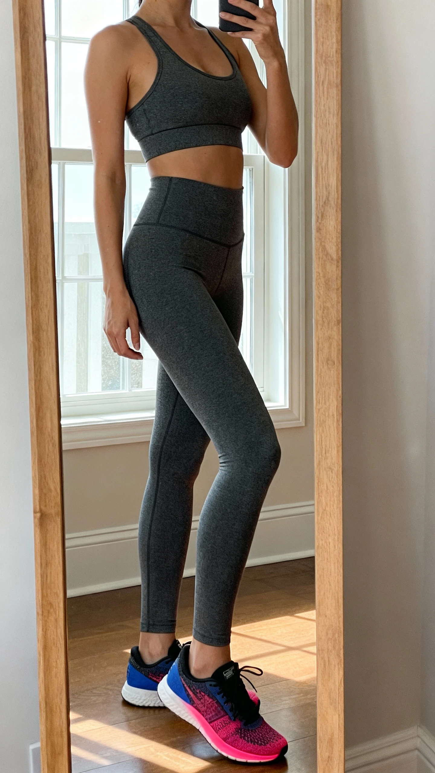 iPhone side-angle mirror selfie of a woman wearing a monochrome charcoal set (high-waisted leggings and matching sports bra) with one intentional pop of color in sneakers, face not visible, cropped at neck, full-length mirror, natural window light, iPhone photo quality