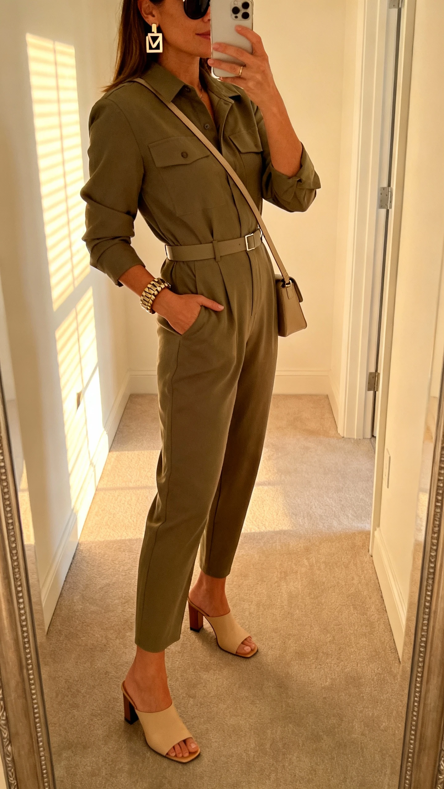 iPhone side-angle selfie of a woman in an olive tailored utility jumpsuit cinched with a minimal belt, heeled mules, geometric earrings, chunky bracelet, medium crossbody, classic sunglasses, face not visible, hallway mirror, late afternoon window light, iPhone photo quality.
