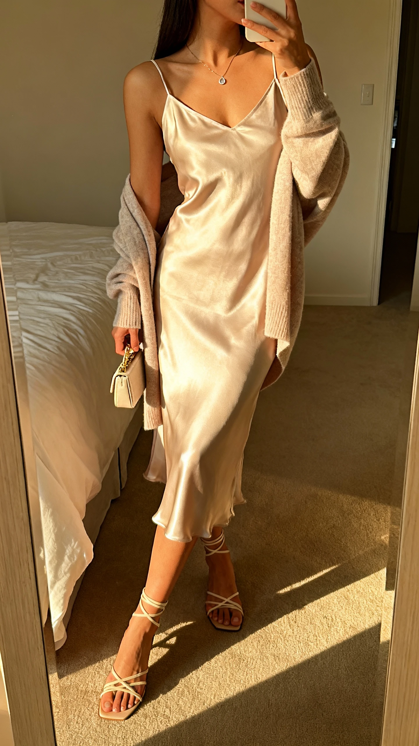 iPhone side-angle selfie of a woman wearing a satin slip dress with a soft cardigan draped over shoulders, strappy sandals, petite clutch, dainty pendant, face not visible, bedroom full-length mirror, golden hour light, iPhone photo quality.